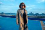 Atriz caracterizada como Odete Roitman usa trench coat bege, óculos escuros e bolsa estruturada enquanto posa em um terraço com vista para o Pão de Açúcar e o mar do Rio de Janeiro ao fundo, no remake da novela Vale Tudo.