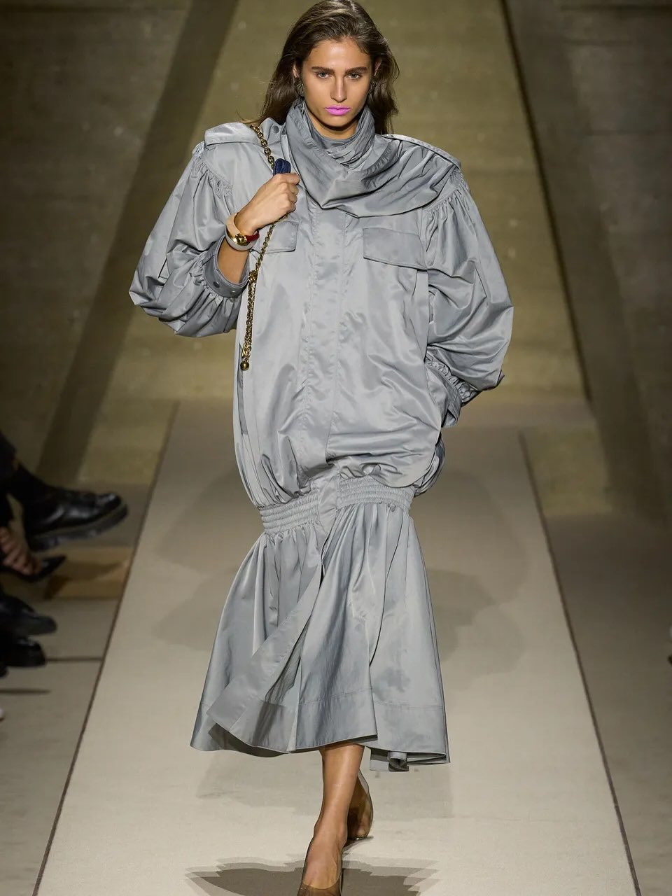 Imagem mostra produção de roupa e maquiagem do desfile da Chloé Primavera/Verão 2026. Mulher morena está de batom rosa-chiclete, cabelos para trás e um vestido longo azul acizentado.