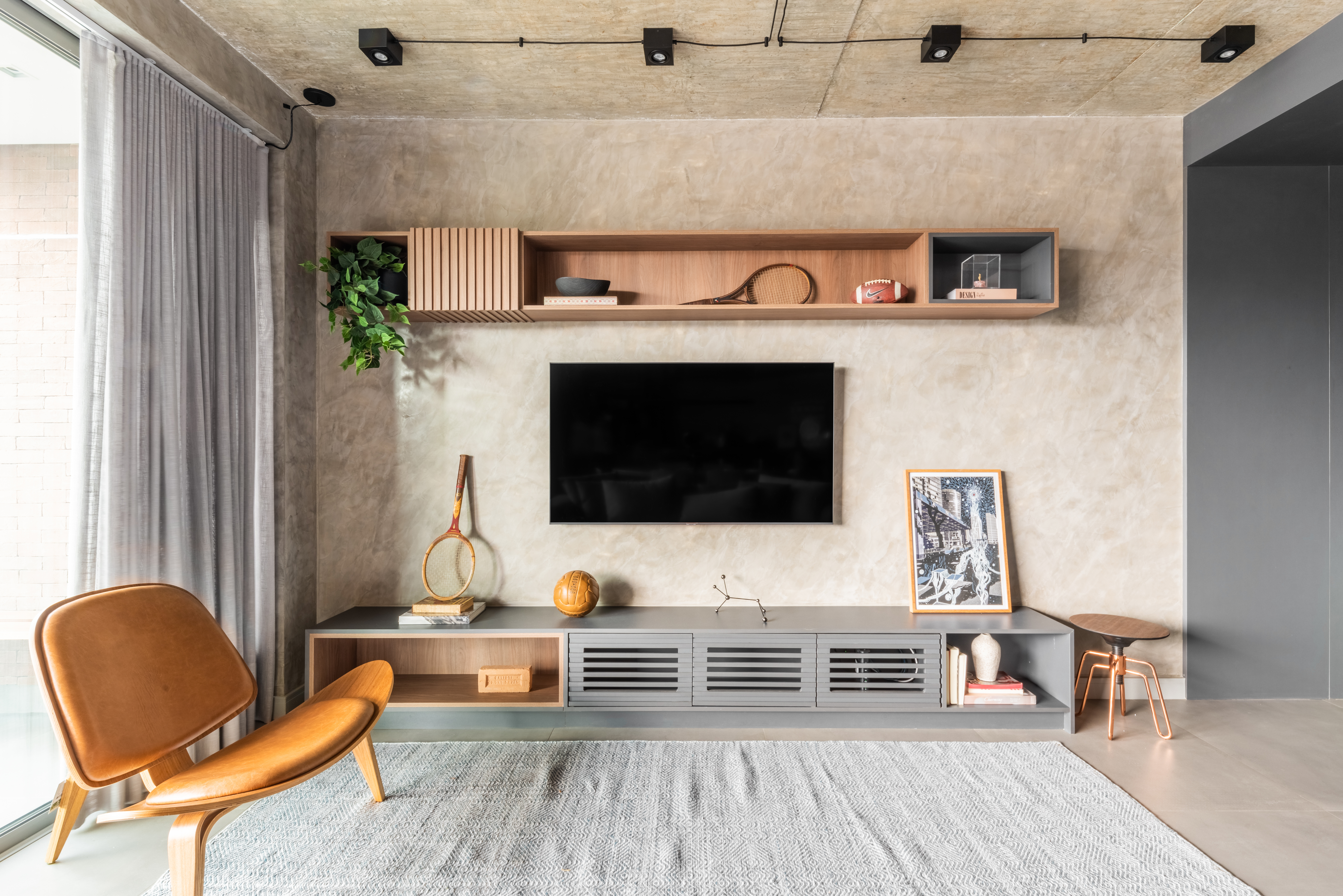 Painel de TV em textura de cimento queimado acompanhado por rack cinza com nichos abertos e objetos decorativos como raquetes vintage, bola de madeira, quadro apoiado e vasos. Prateleiras superiores exibem pe&ccedil;as em madeira e cer&acirc;mica, enquanto uma cadeira de couro marrom comp&otilde;e o ambiente industrial acolhedor