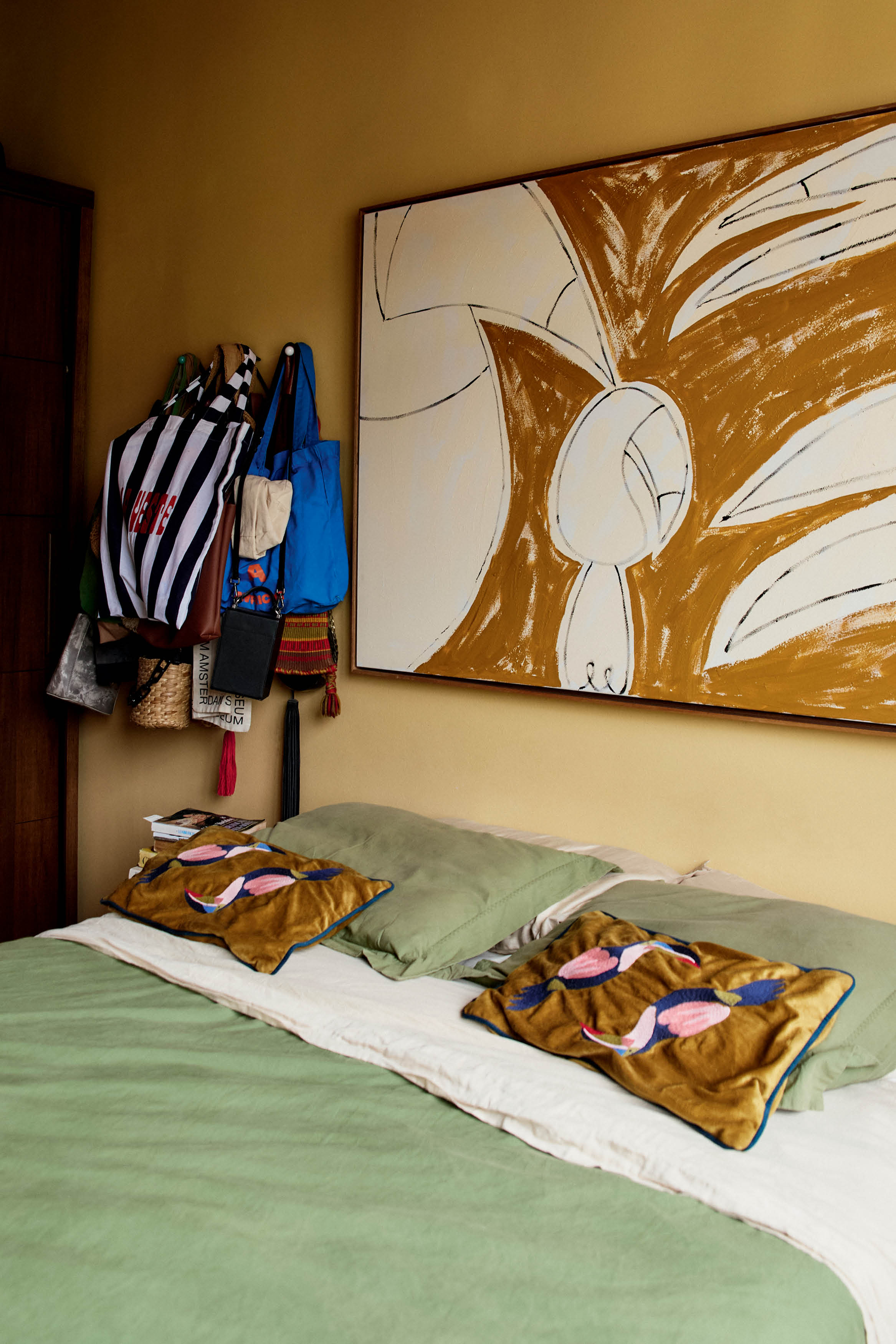 Quarto com parede amarela, quadro e casa de casal com roupa verde e branca.