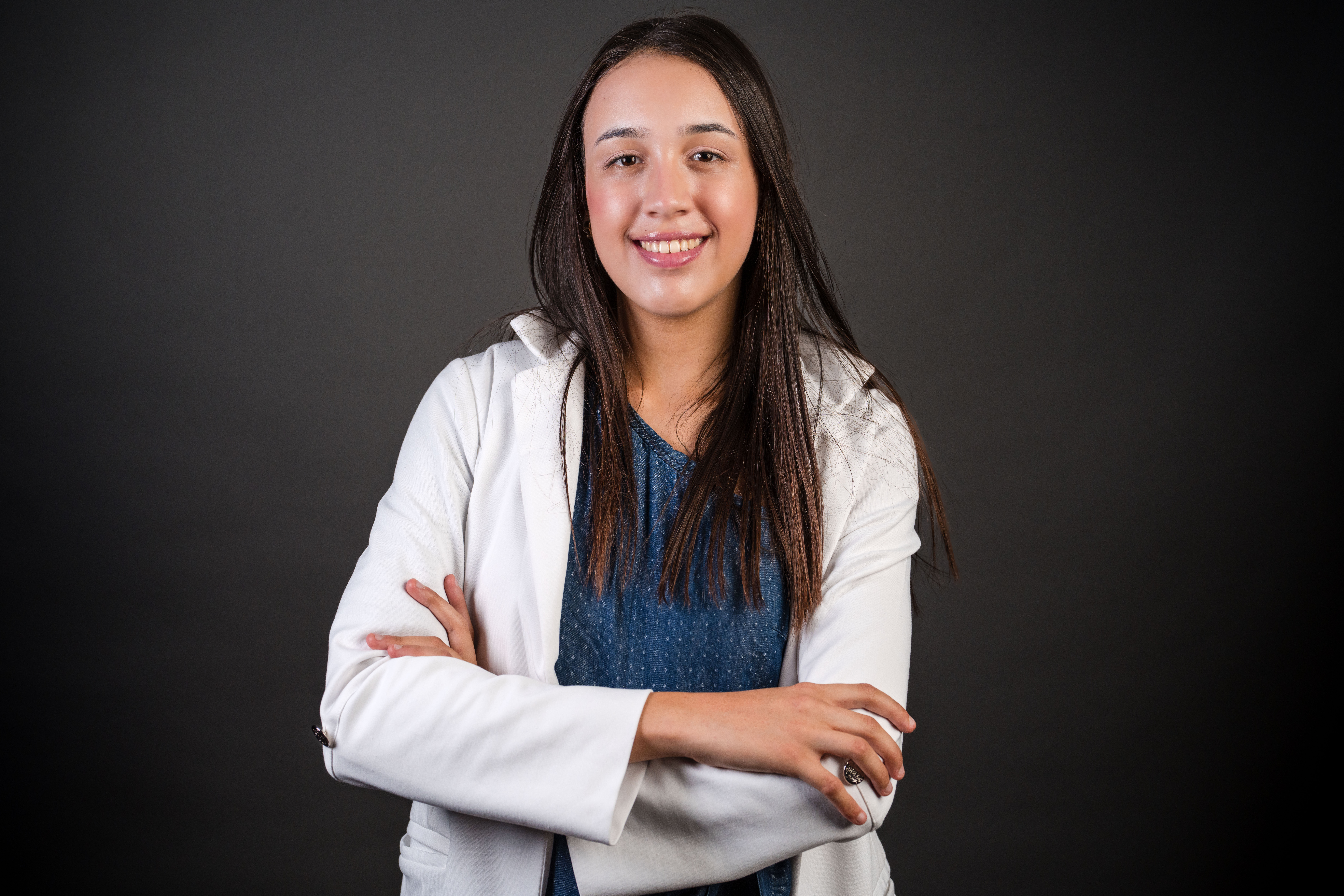 Millena Xavier Martins, destaque brasileiro em competi&ccedil;&otilde;es cient&iacute;ficas e premiada pela Forbes Under 30, posa sorridente com jaleco branco e blusa azul, em est&uacute;dio com fundo neutro. A fotografia evidencia sua postura segura e seu papel como jovem refer&ecirc;ncia na &aacute;rea de intelig&ecirc;ncia artificial e educa&ccedil;&atilde;o