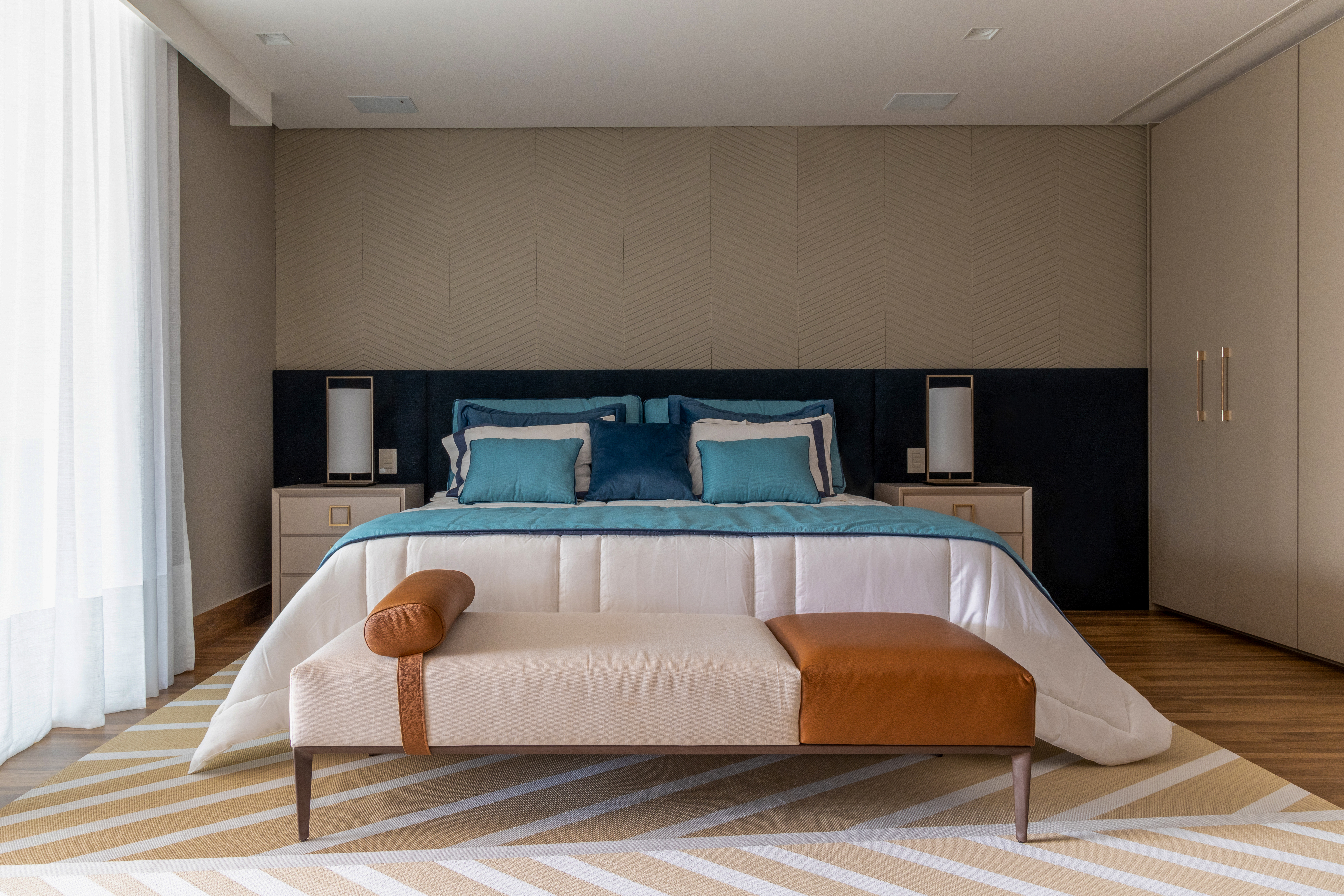 Quarto de casal com cama king e roupa de cama branca e e em tons de azul. M&oacute;vel de apoio de cada lado do casal e banco em frente &agrave; cama.