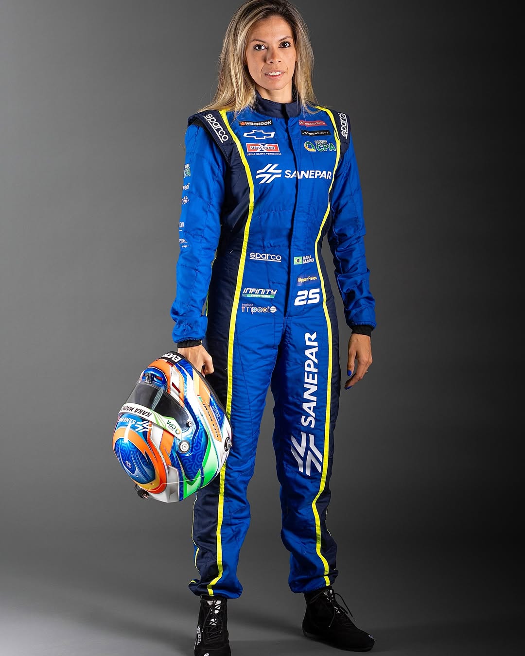 Mulher loira posando para foto em est&uacute;dio usando roupa de pilota e segurando capacete na m&atilde;o.