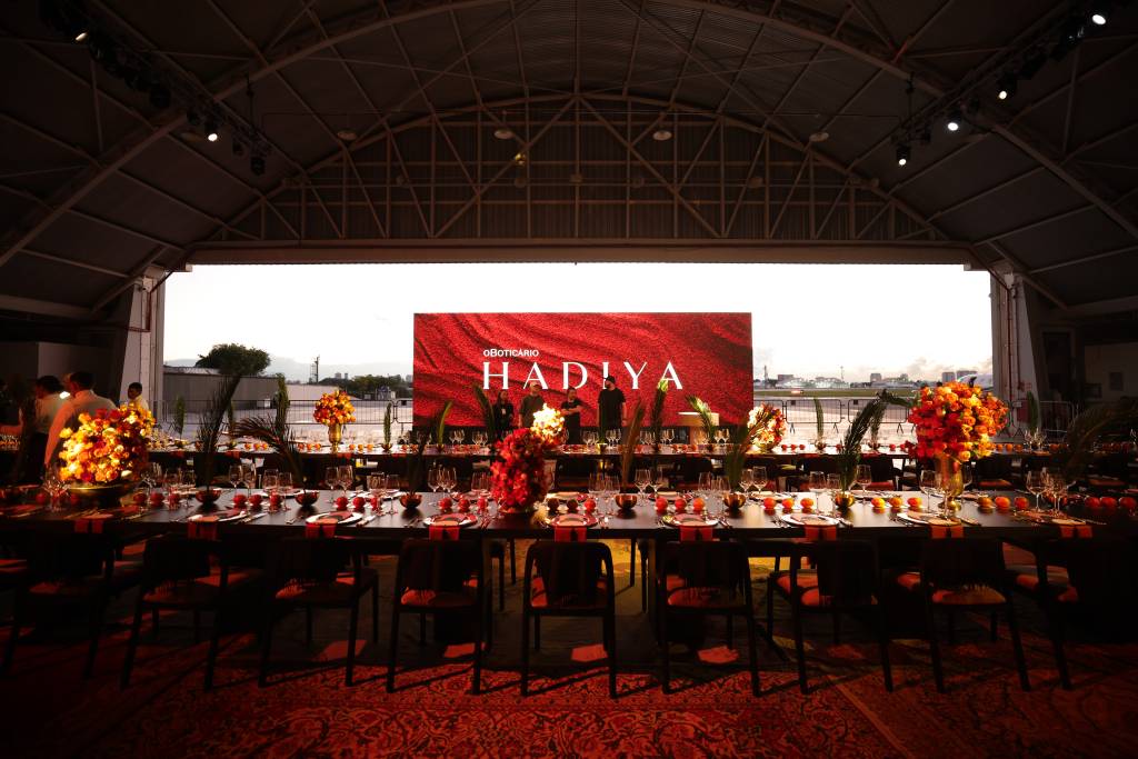 Ambiente do evento de lançamento de Hadiya em São Paulo, com mesas decoradas em tons quentes, arranjos florais e painel ao fundo com o nome do perfume. Pessoas circulam pelo local, incluindo convidados como Silvia Braz, o perfumista Adilson Rato e a VP de marketing Renata Gomide.