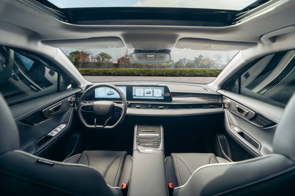 Interior do Omoda 5 visto a partir do banco traseiro, mostrando o painel digital widescreen, volante multifuncional, acabamento em tons escuros e teto panorâmico, com destaque para o design moderno e minimalista do SUV