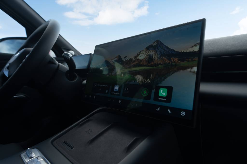 Interior do Omoda 7 mostrando a grande tela multimídia UHD de 15,6 polegadas, posicionada no centro do painel, exibindo navegação, telefone e configurações, com volante e detalhes do cockpit ao fundo