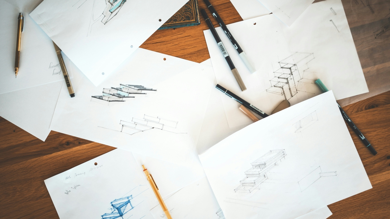 Papéis com desenhos rascunhados e canetas espalhados sobre mesa