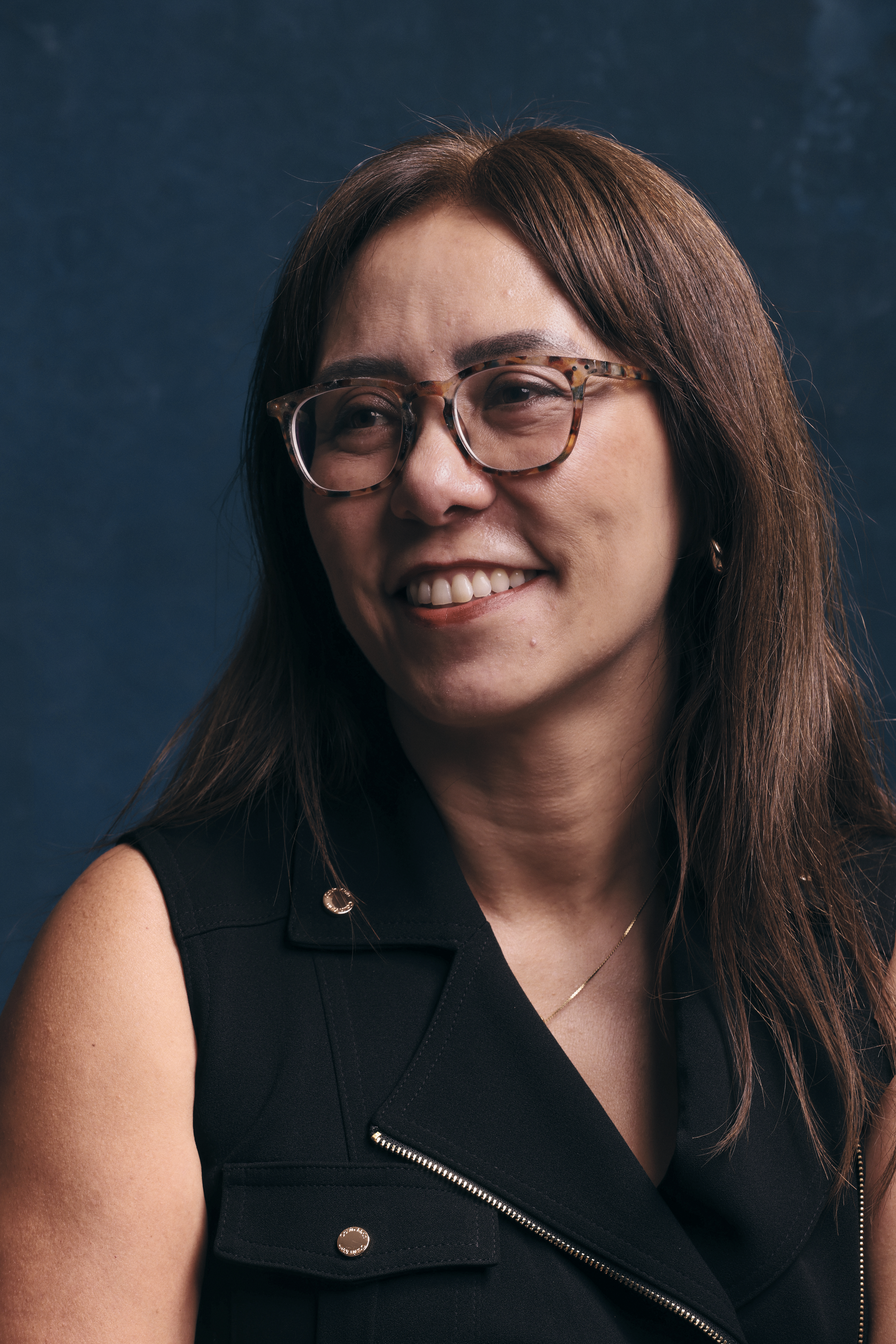 Foto de Claudia Suemoto. Ela &eacute; amarela, tem cabelo castanho e liso e est&aacute; sentada. Usa um colete azul marinho e, atr&aacute;s dela, h&aacute; tamb&eacute;m um cen&aacute;rio azul marinho. A imagem foca em seu rosto