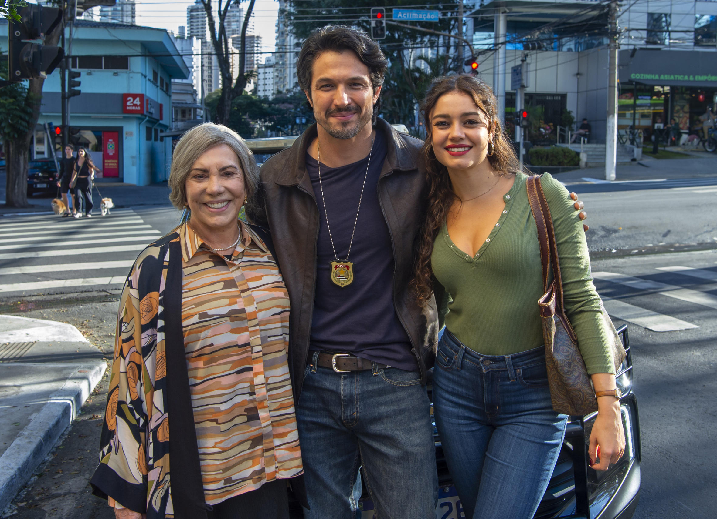 Três pessoas do elenco de Três Graças (Arlete Salles, Romulo Estrela e Sophie Charlotte) posam juntas em uma rua urbana, em frente a uma faixa de pedestres. Um homem ao centro usa jaqueta escura e exibe um distintivo pendurado no pescoço, enquanto duas mulheres, uma mais velha à esquerda e outra mais jovem à direita, sorriem ao seu lado. Ao fundo, aparecem prédios, semáforos e pedestres, compondo um cenário de cidade em movimento.