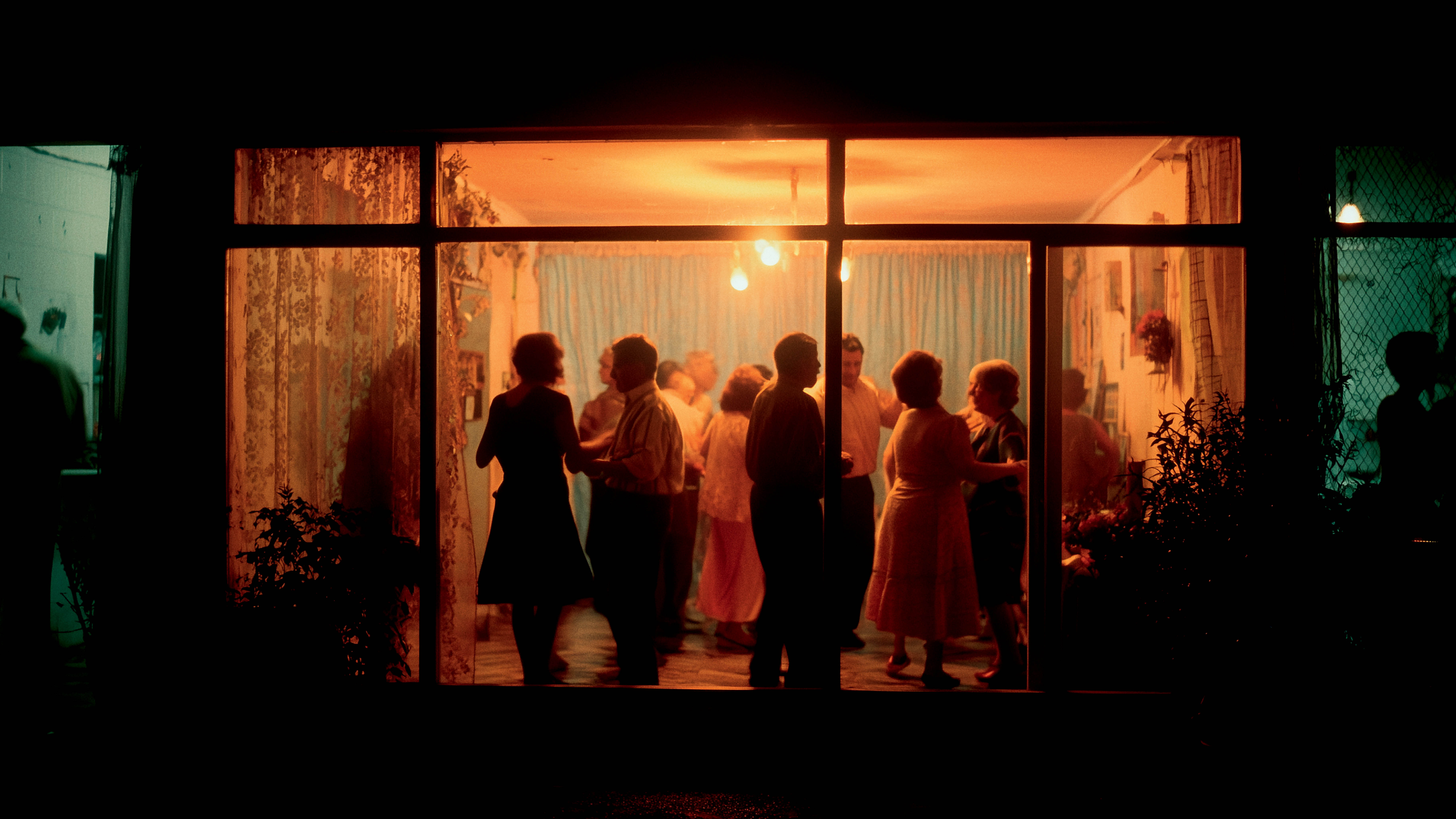 Pessoas dançam em uma festa vista por uma janela iluminada, com silhuetas em movimento; imagem simboliza convivência comunitária, socialização e combate à solidão na terceira idade