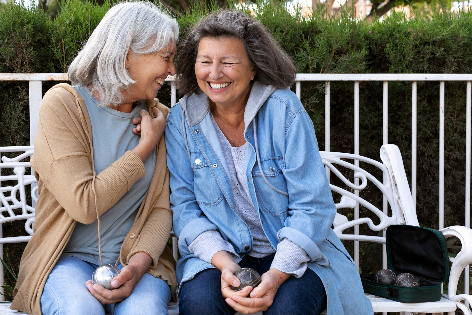 Duas mulheres idosas sorriem e conversam sentadas em um banco ao ar livre, segurando bolas de bocha; cena representa amizade na terceira idade e envelhecimento com apoio social