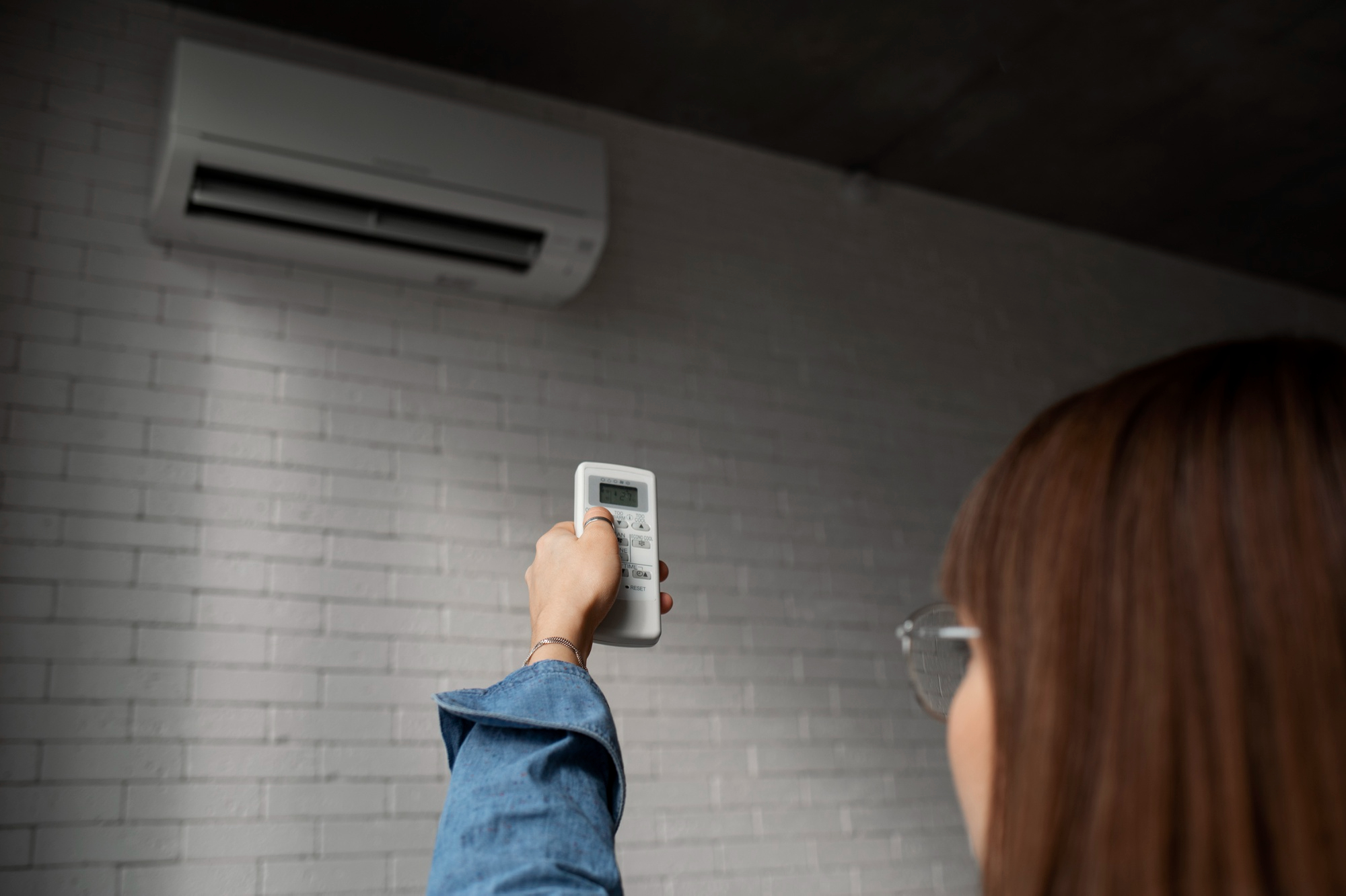 Pessoa de perfil usando controle remoto para ligar o ar-condicionado split instalado na parede de tijolinhos claros, em casa, ilustrando dicas para economizar energia e reduzir a conta de luz no verão.