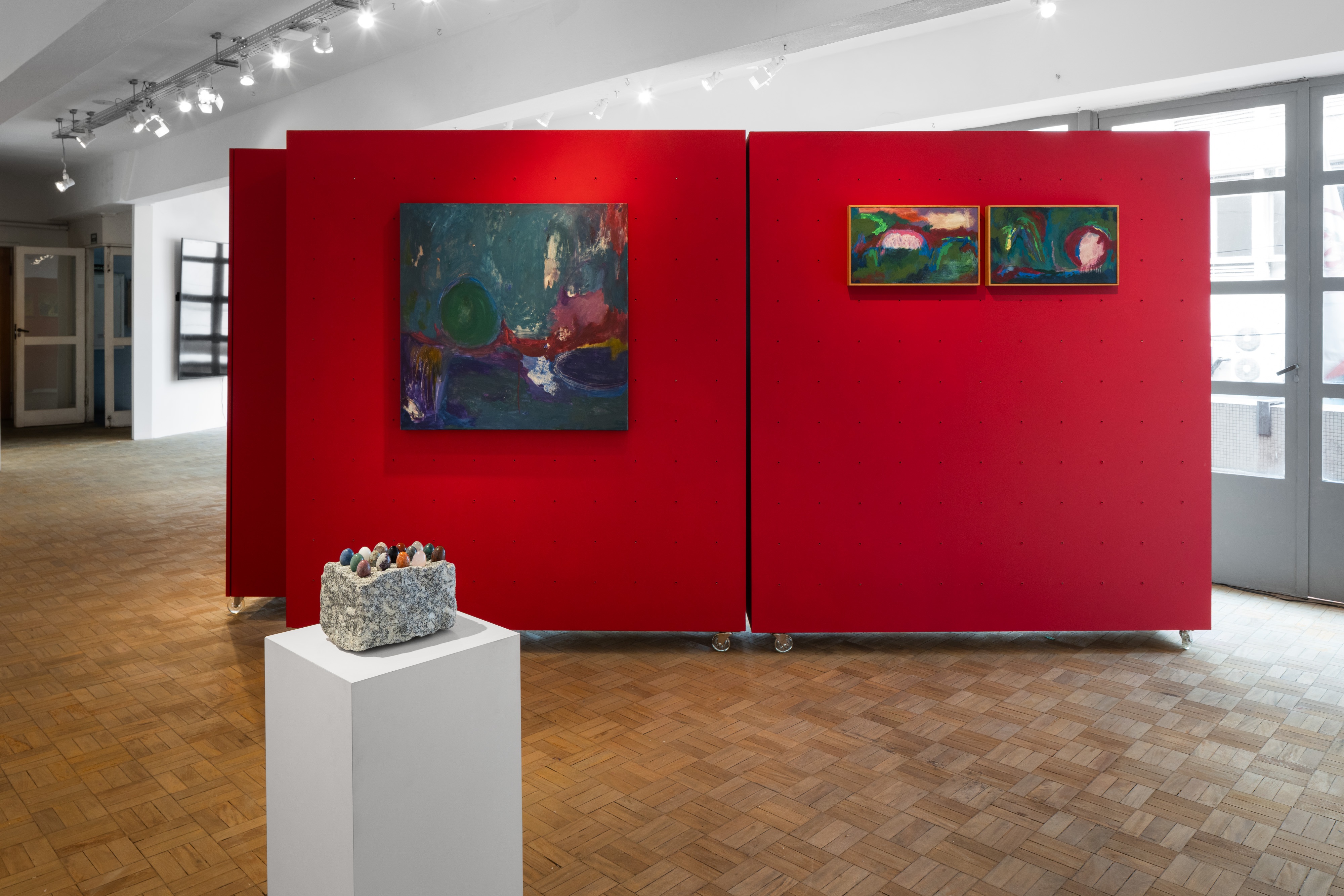 Vista interna da WG Galeria, no centro de São Paulo, com obras de arte expostas em painéis vermelhos e escultura sobre pedestal durante a exposição Um delírio de carnaval.