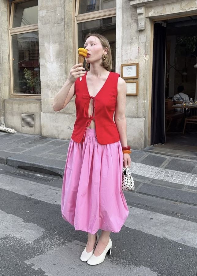 Foto de uma mulher branca usando um look composto por colete vermelho, saia rosa e bolsa pequena