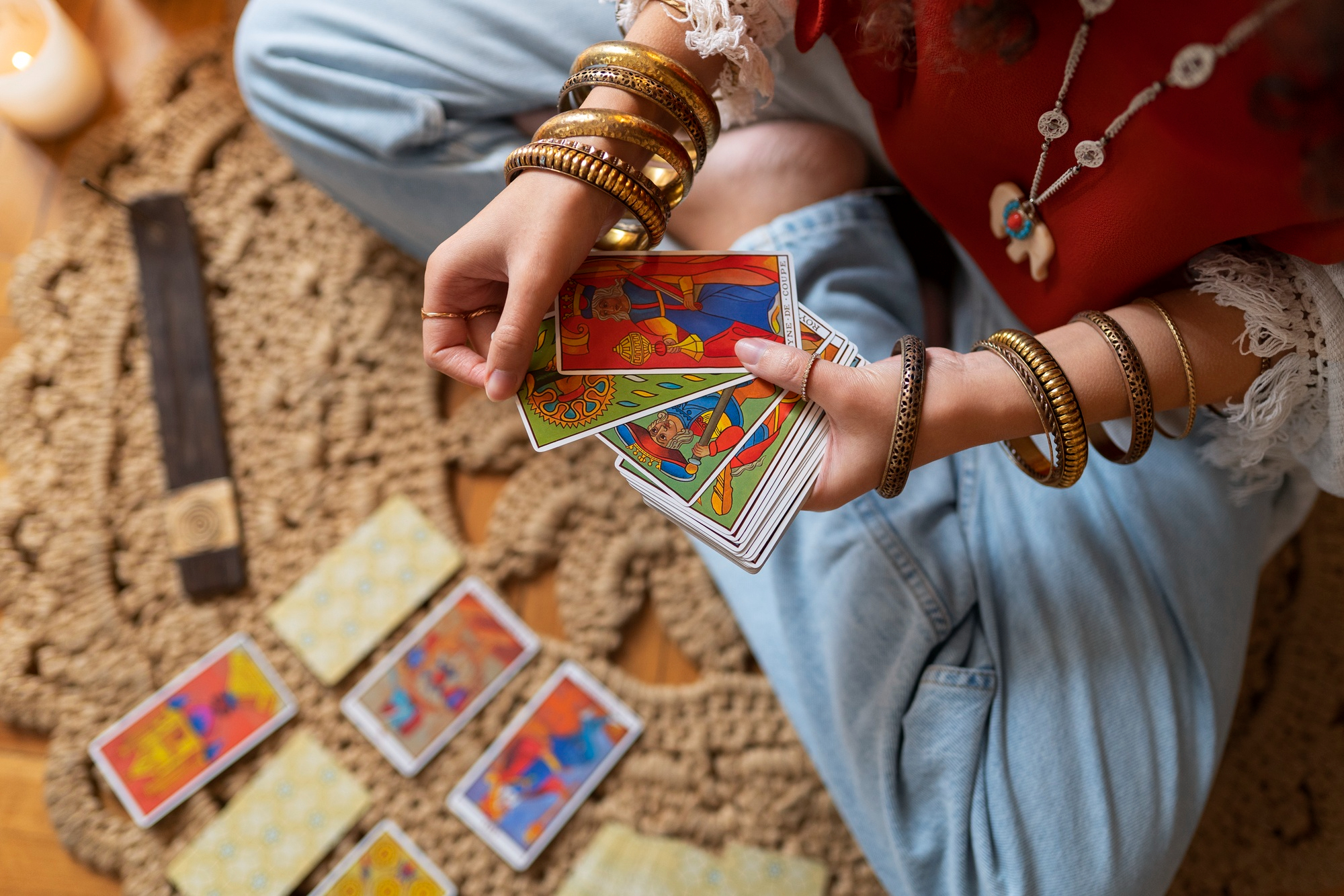 Mãos segurando cartas de Tarô durante leitura sobre o Cinco de Ouros, com baralho espalhado no chão e velas ao fundo, simbolizando reflexão sobre dinheiro, autoestima, limites e superação de dificuldades em janeiro de 2026