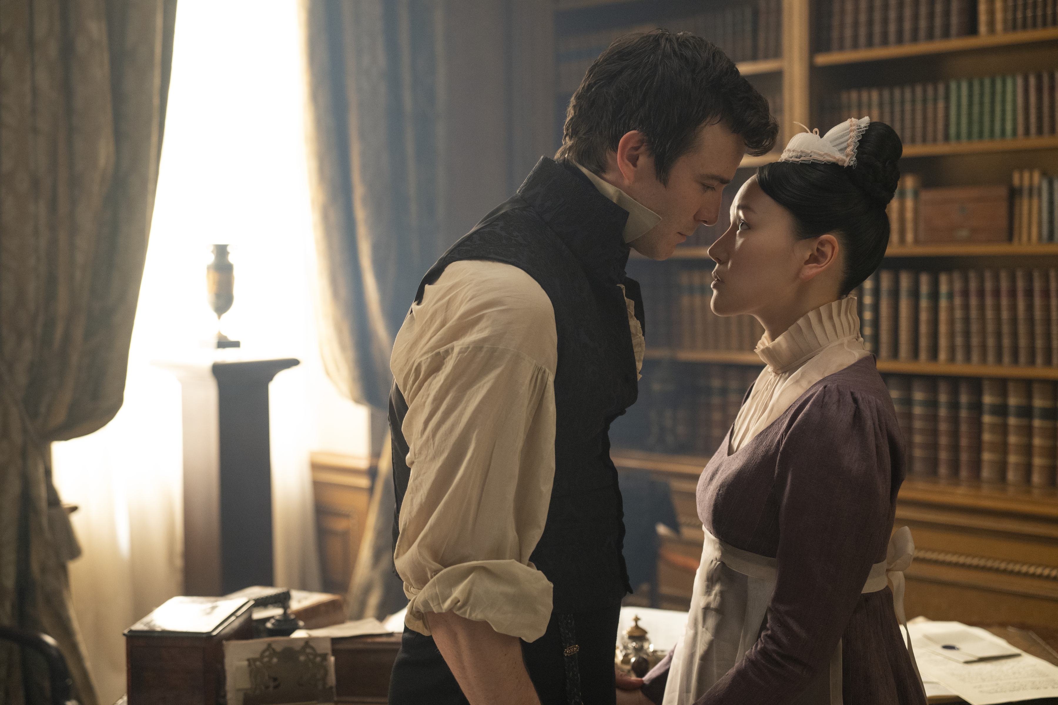 O casal se aproxima com delicadeza em meio a estantes de livros, refor&ccedil;ando o romantismo e a tens&atilde;o crescente entre os dois.