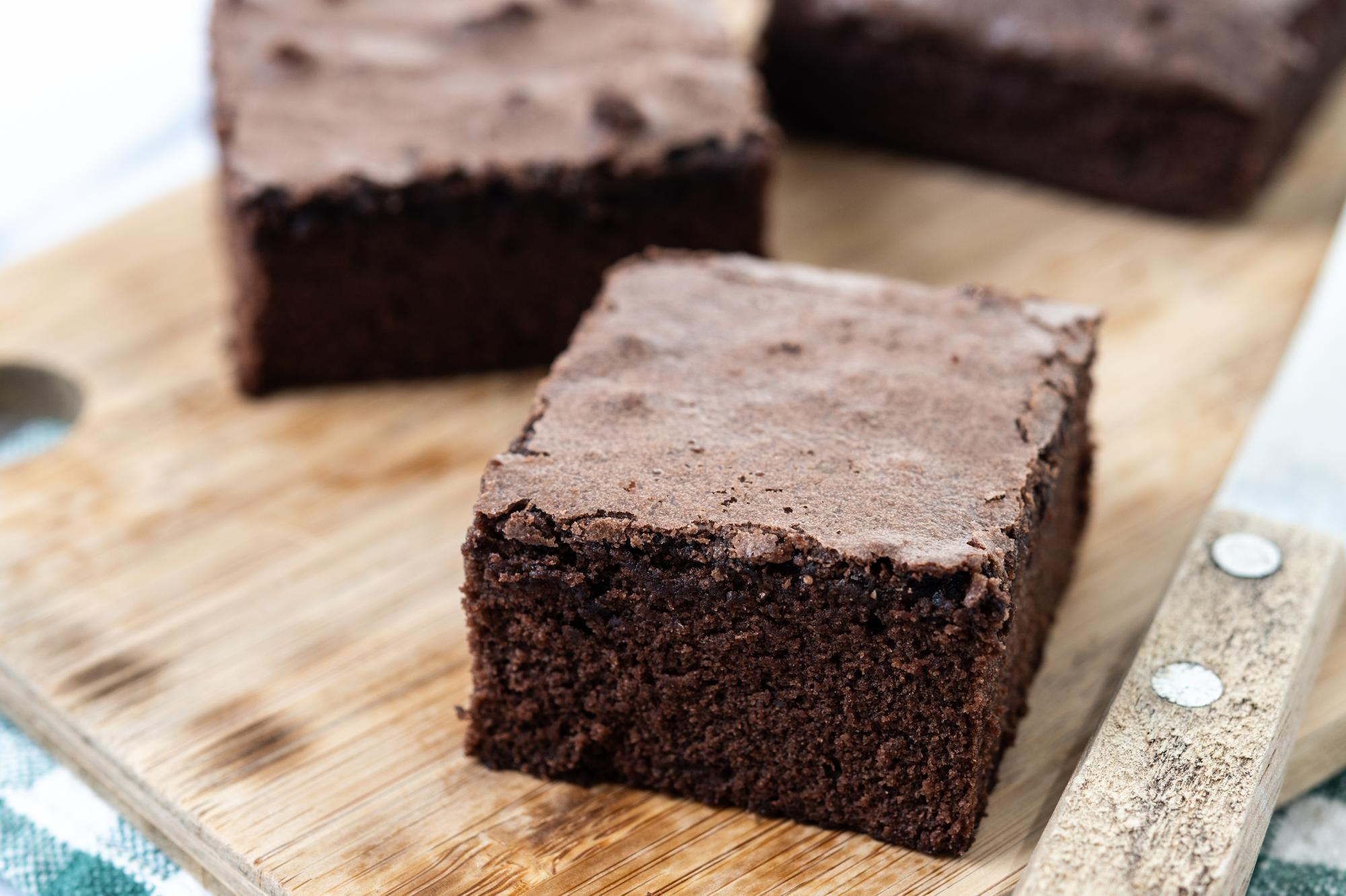 Peda&ccedil;os grossos de brownie com textura densa e escura, sobre uma t&aacute;bua de madeira. Um peda&ccedil;o aparece em destaque na frente, enquanto outros ficam desfocados ao fundo