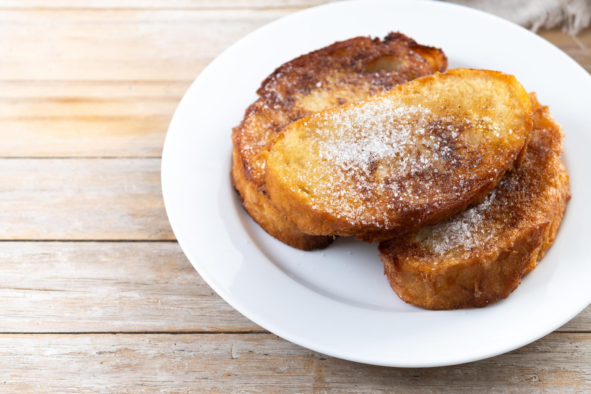 Tr&ecirc;s fatias de rabanada douradas e a&ccedil;ucaradas empilhadas em um prato branco, com a&ccedil;&uacute;car cristal vis&iacute;vel na superf&iacute;cie, sobre uma mesa de madeira clara.