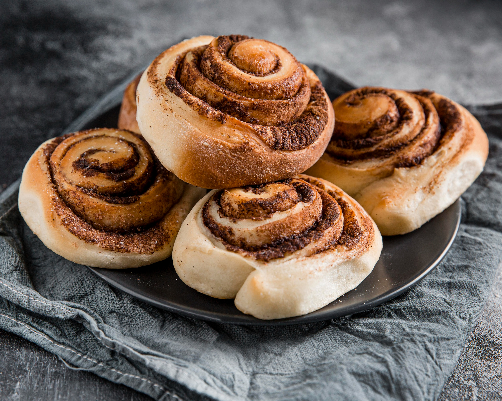 Quatro cinnamon rolls assados e dourados, com espiral vis&iacute;vel de canela e a&ccedil;&uacute;car, empilhados em um prato escuro sobre um pano cinza, criando uma composi&ccedil;&atilde;o r&uacute;stica e aconchegante.