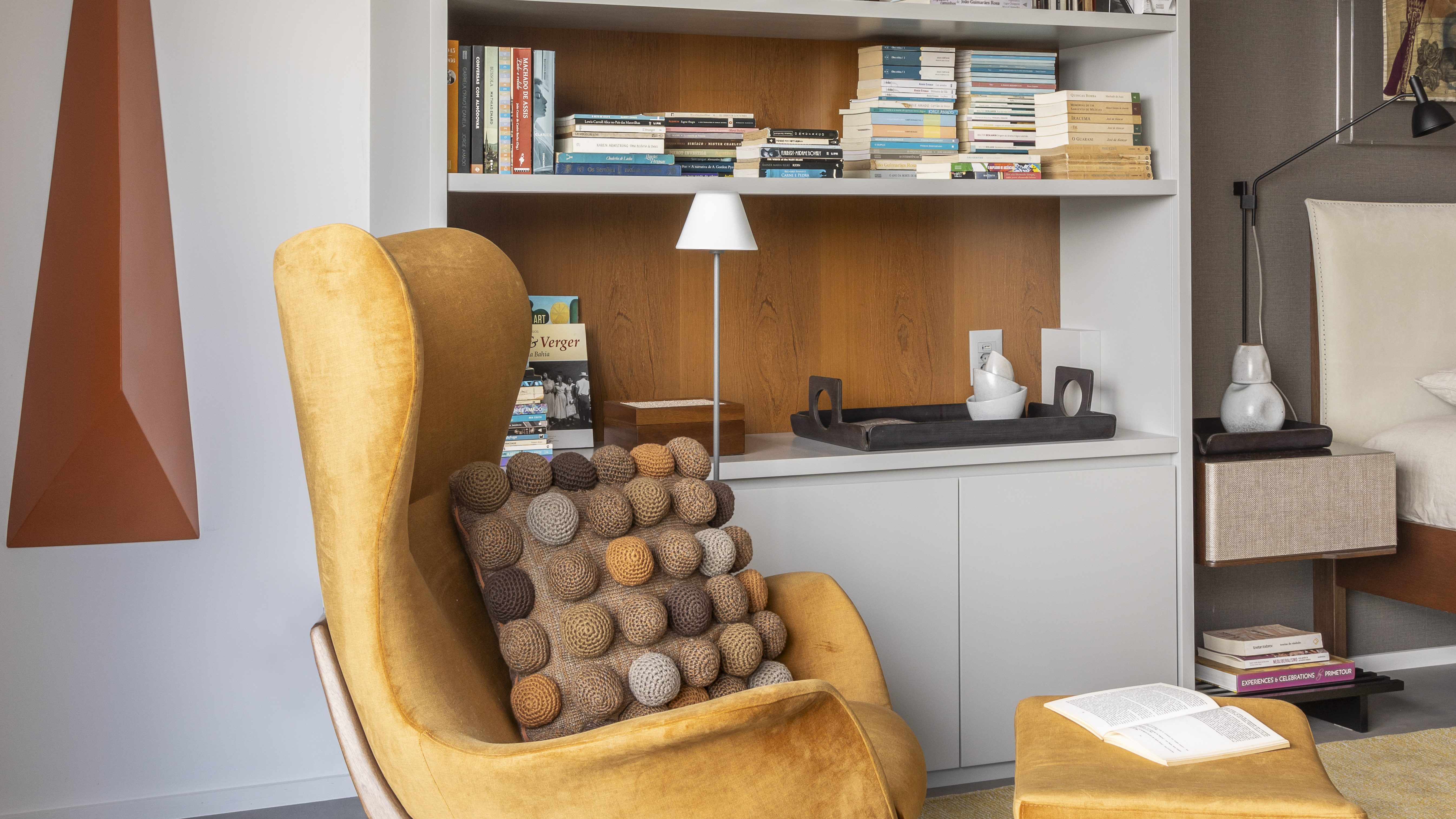 Cantinho da leitura com poltrona amarelo mostarde e prateleira de livros atrás