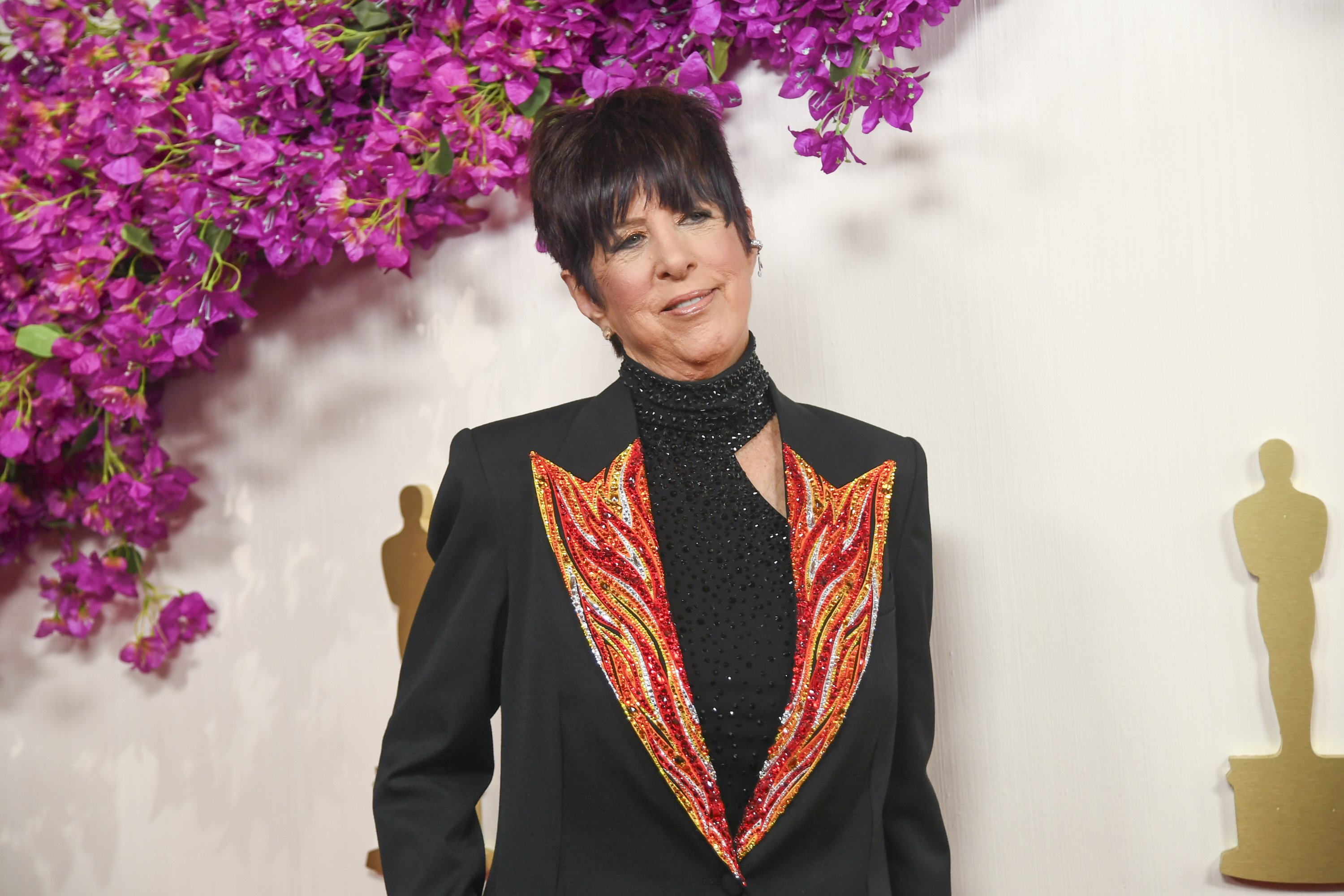 Diane Warren posa diante de um cen&aacute;rio decorado com flores rosas e silhuetas douradas da estatueta do Oscar. Vestindo um blazer preto com detalhes em vermelho e laranja que lembram chamas, a compositora sorri levemente para os fot&oacute;grafos.