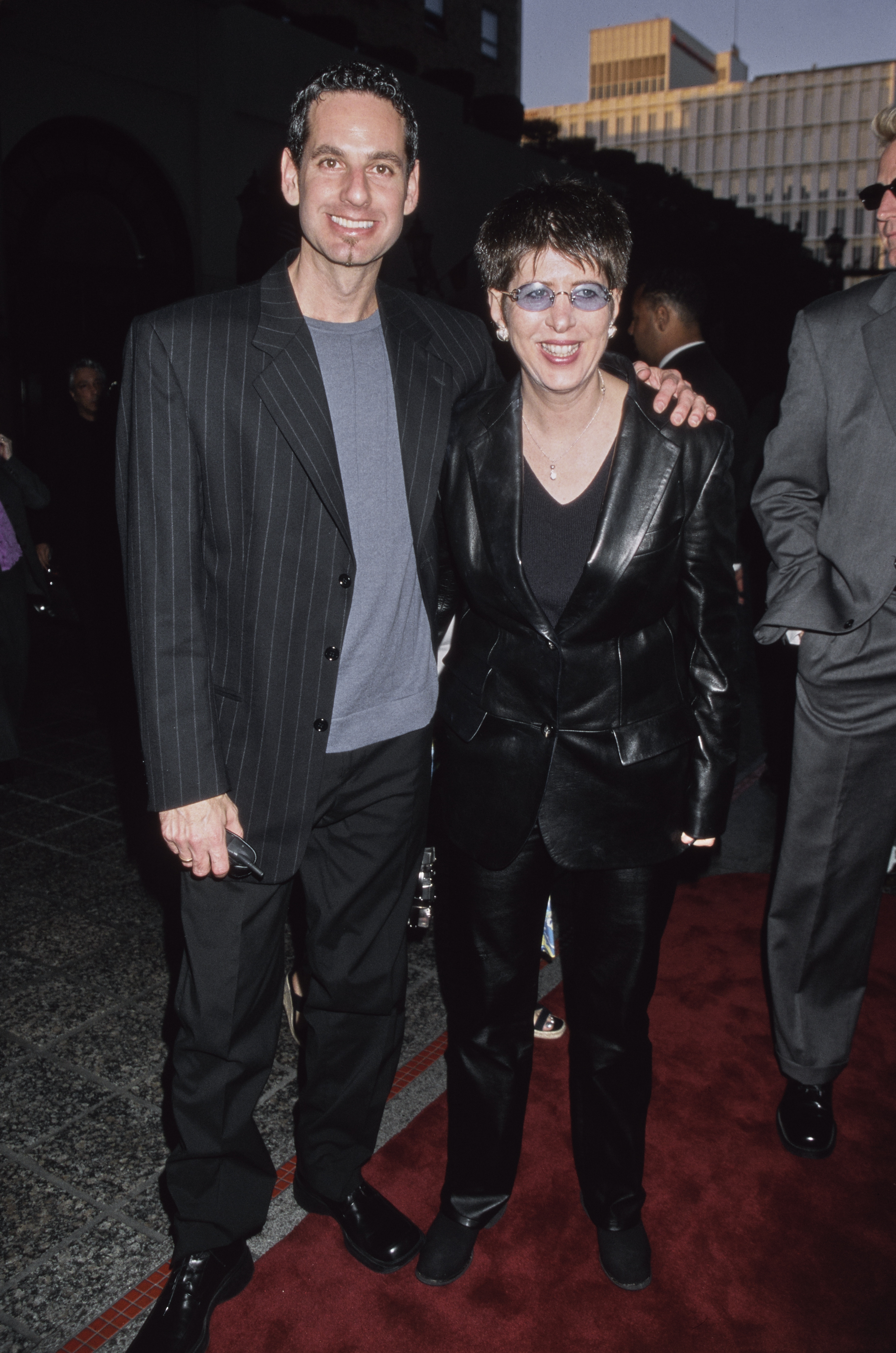 Registro de arquivo mostra a compositora Diane Warren ao lado de um acompanhante durante um evento de tapete vermelho. Ela aparece usando &oacute;culos escuros de lentes claras e um conjunto preto de couro, enquanto ambos sorriem para as c&acirc;meras. A cena tem o clima t&iacute;pico das estreias e premia&ccedil;&otilde;es de Hollywood.