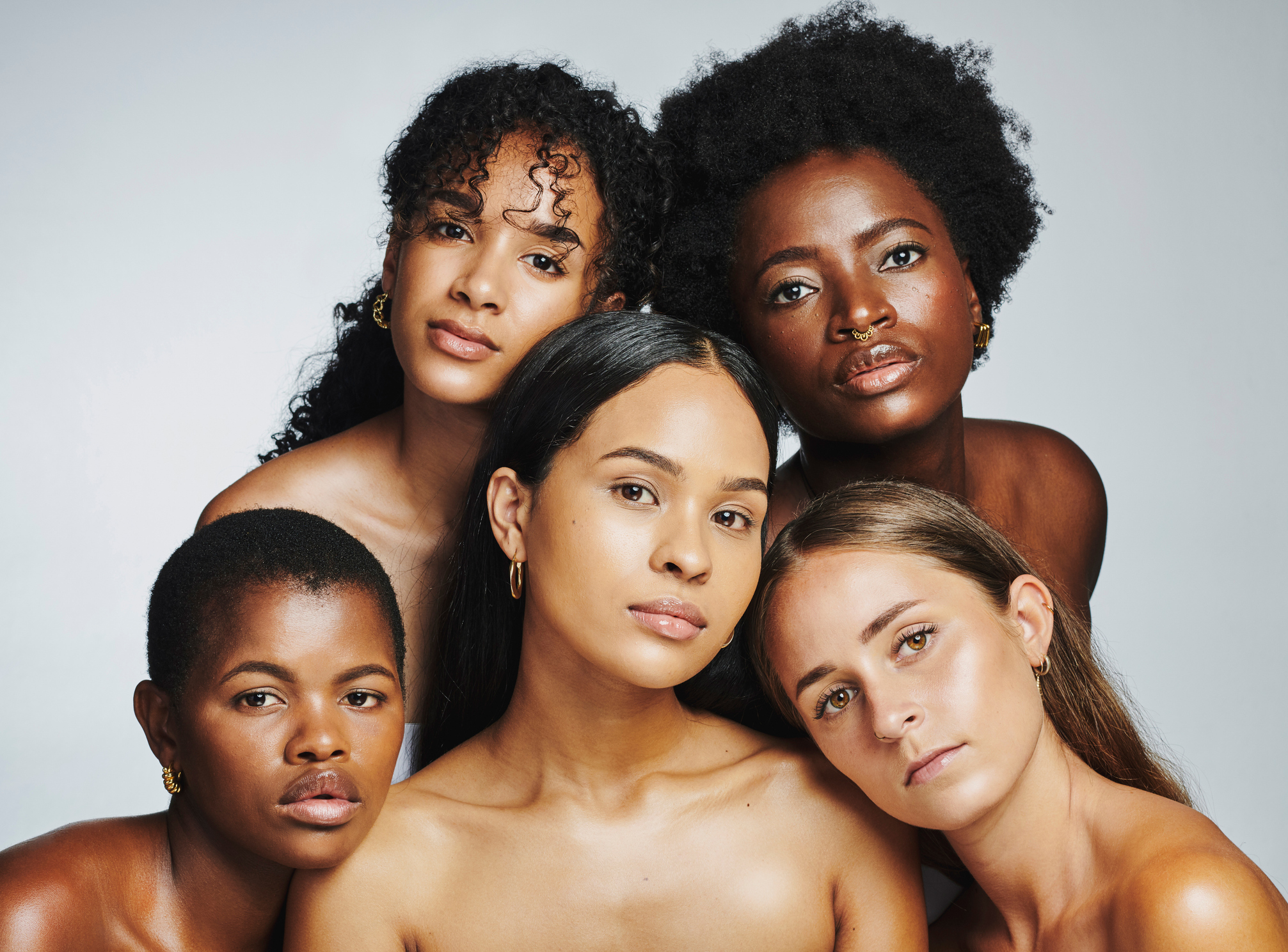 Cinco mulheres com diferentes tons de pele posam juntas, destacando a diversidade de rostos e características em um retrato de beleza natural.