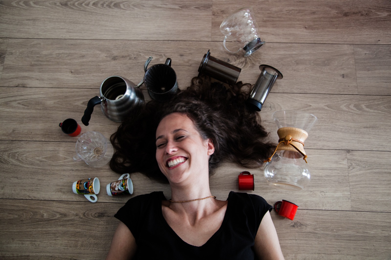 Kelly Stein sorrindo deitada no ch&atilde;o cercada por utens&iacute;lios de caf&eacute;, como cafeteiras e x&iacute;caras, imagem que representa pausa, rotina e reconex&atilde;o com si mesma no meio do cotidiano