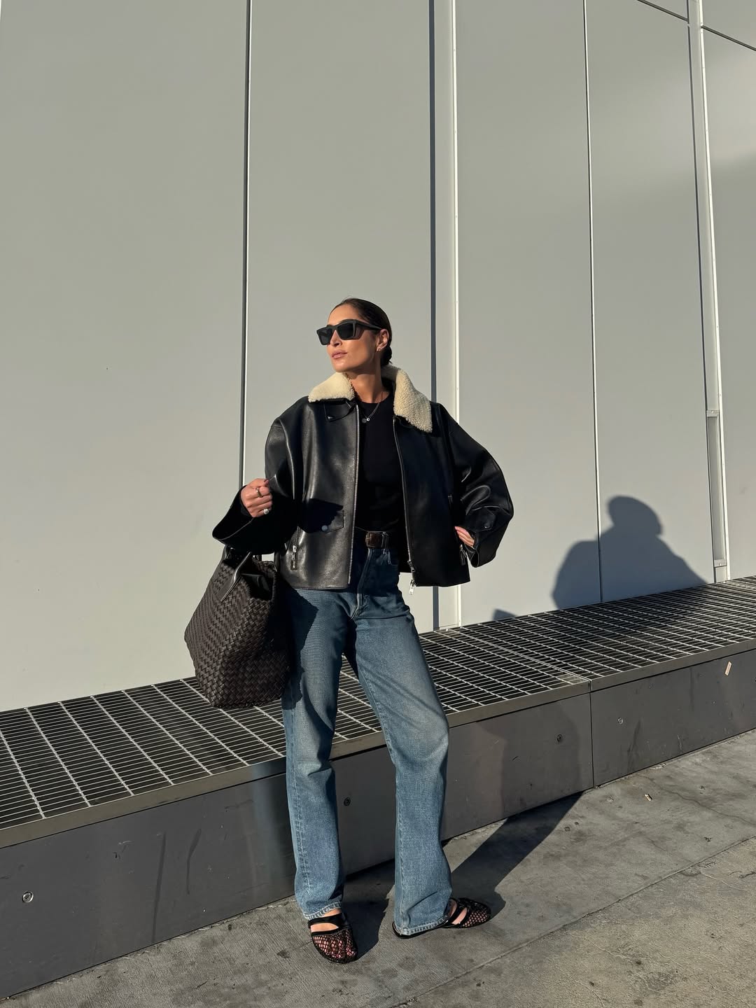 Foto de uma mulher morena usando um look composto por jaqueta de couro, cal&ccedil;a boot cut, bolsa preta, &oacute;culos de sol e sapatilha transparente