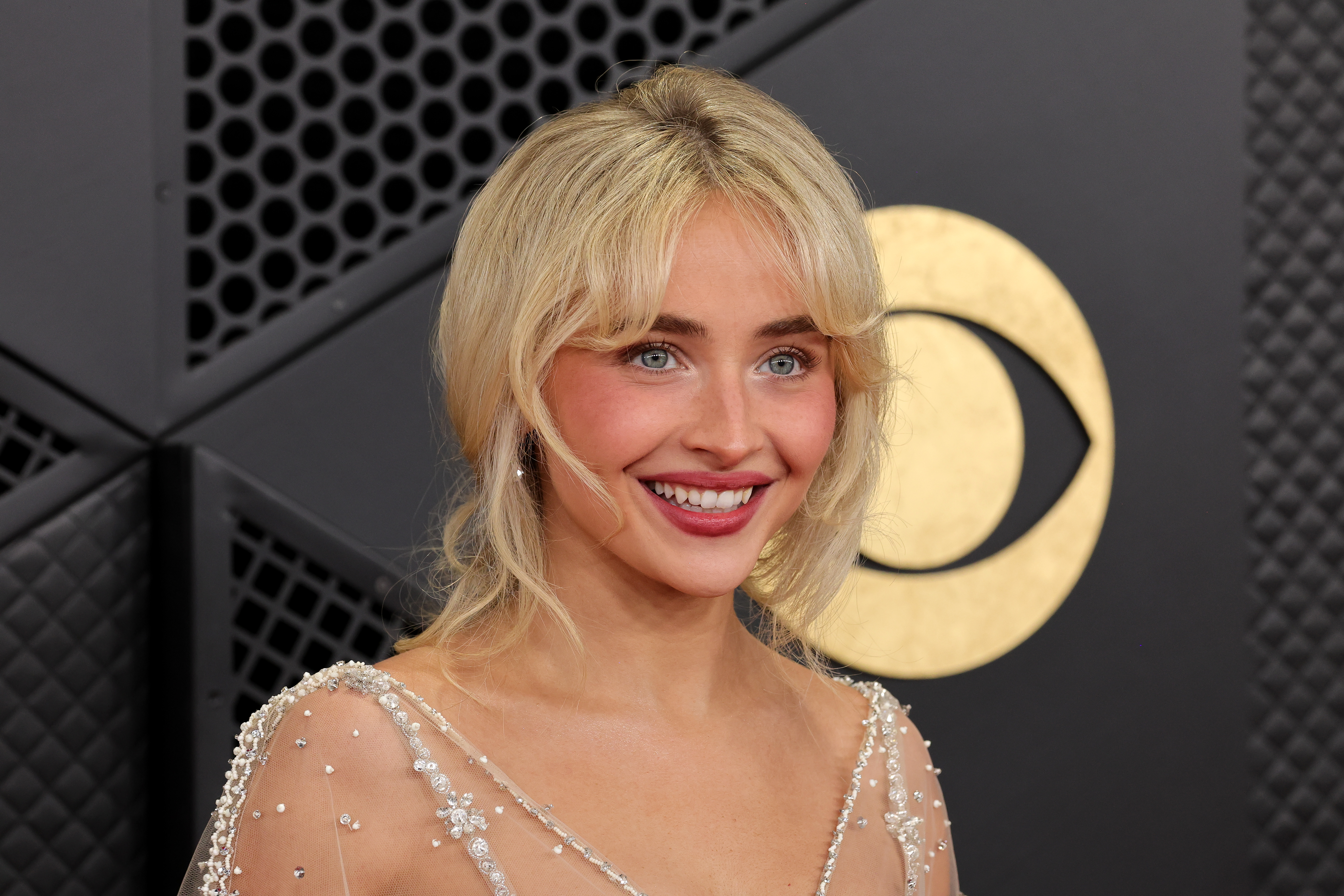 Sabrina Carpenter com maquiagem suave e iluminada, usando blush rosado, pele glow, lábios brilhantes e franja desfiada em evento