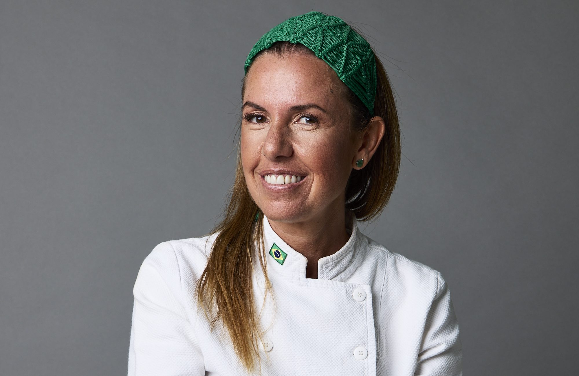 Retrato da chef Morena Leite de dólmã branca e sentada num banco de madeira