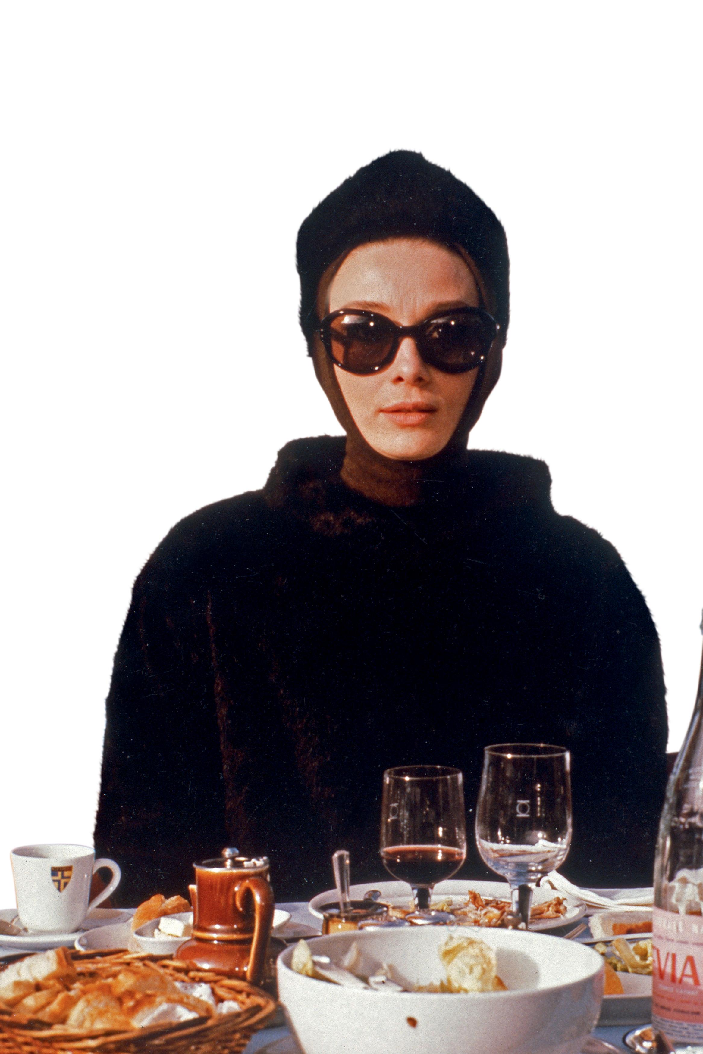 Audrey Hepburn surge em um visual sofisticado, com &oacute;culos escuros e roupa escura elegante, sentada &agrave; mesa durante uma refei&ccedil;&atilde;o. A imagem remete &agrave; aura refinada que marcou sua colabora&ccedil;&atilde;o com Hubert de Givenchy.