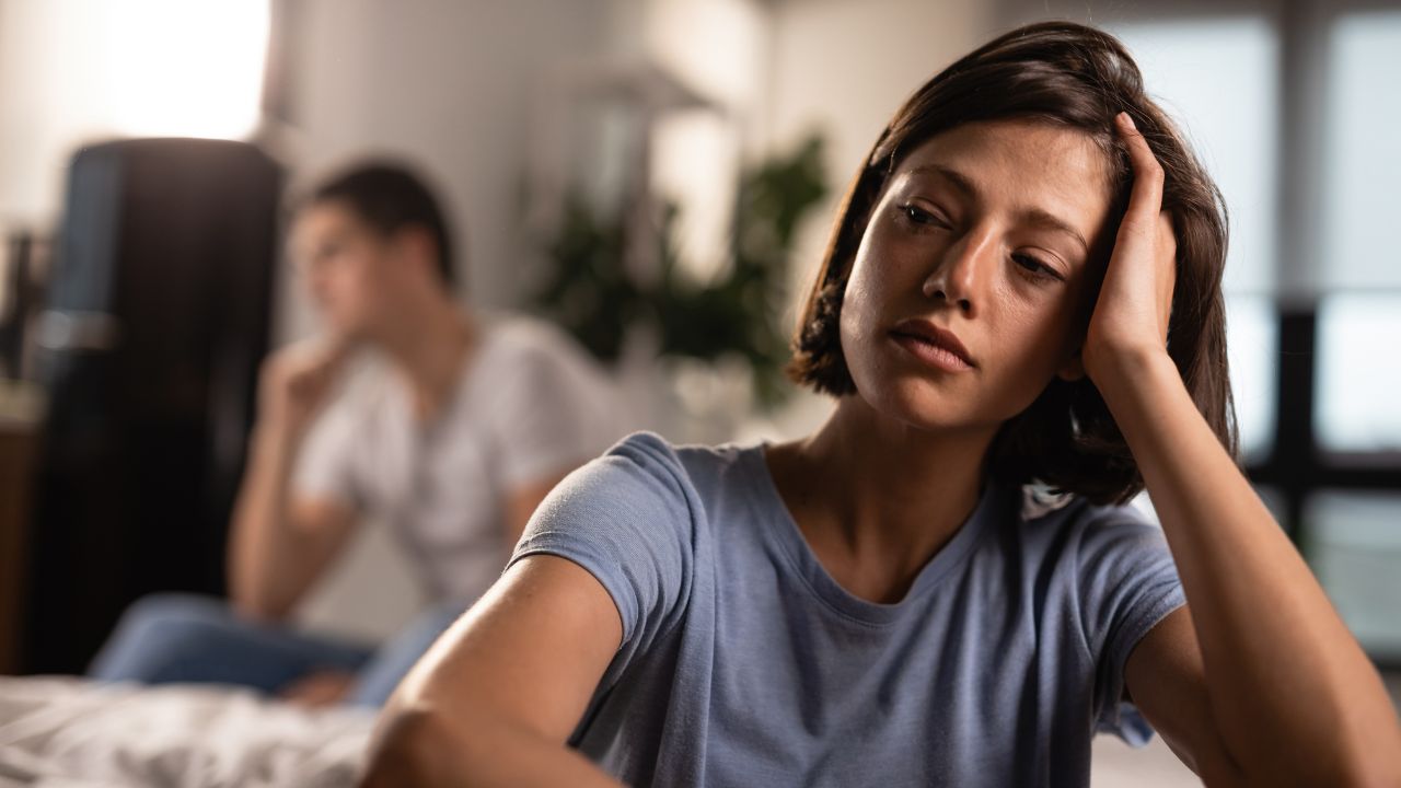 Mulher sentada pensativa com expressão triste e homem sentado ao fundo, também pensativo
