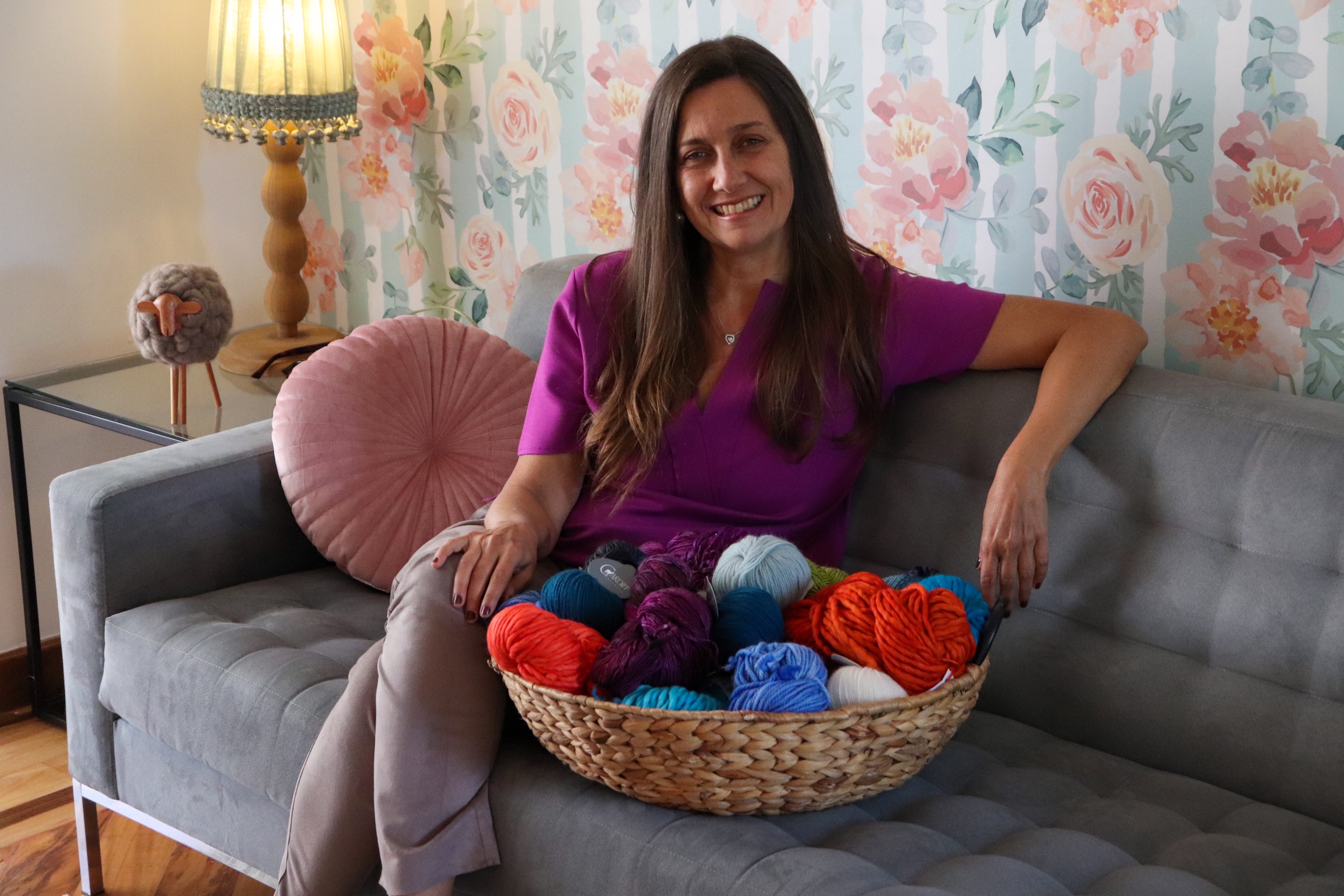 Foto de Aida Fonseca, proprietária da Novelaria. Ela está sentada num sofá cinza e segurando vários fios de tecelagem. Ela tem cabelo grisalho, médio e usa blusa roxa