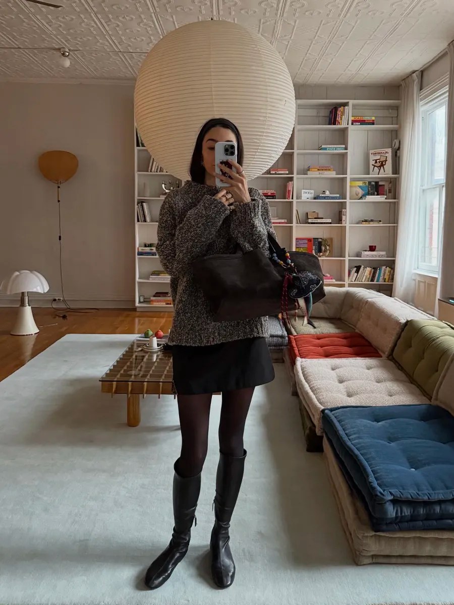 Foto de uma mulher usando um look composto por agasalho cinza, meia-calça, saia e bota preta