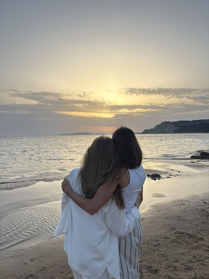 Duas mulheres abraçadas observando o pôr do sol na praia.