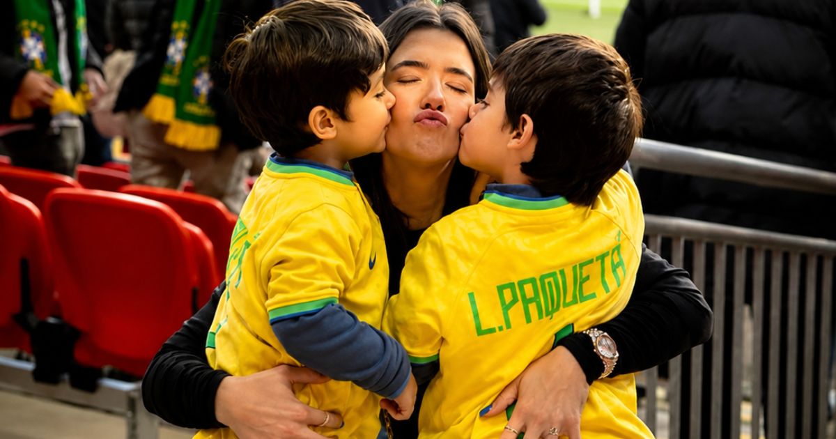 Mulher de cabelos escuros, olhos fechados e boca em bico, recebendo beijos nas bochechas de dois meninos pequenos, ambos vestindo camisas amarelas da sele&ccedil;&atilde;o brasileira com 
