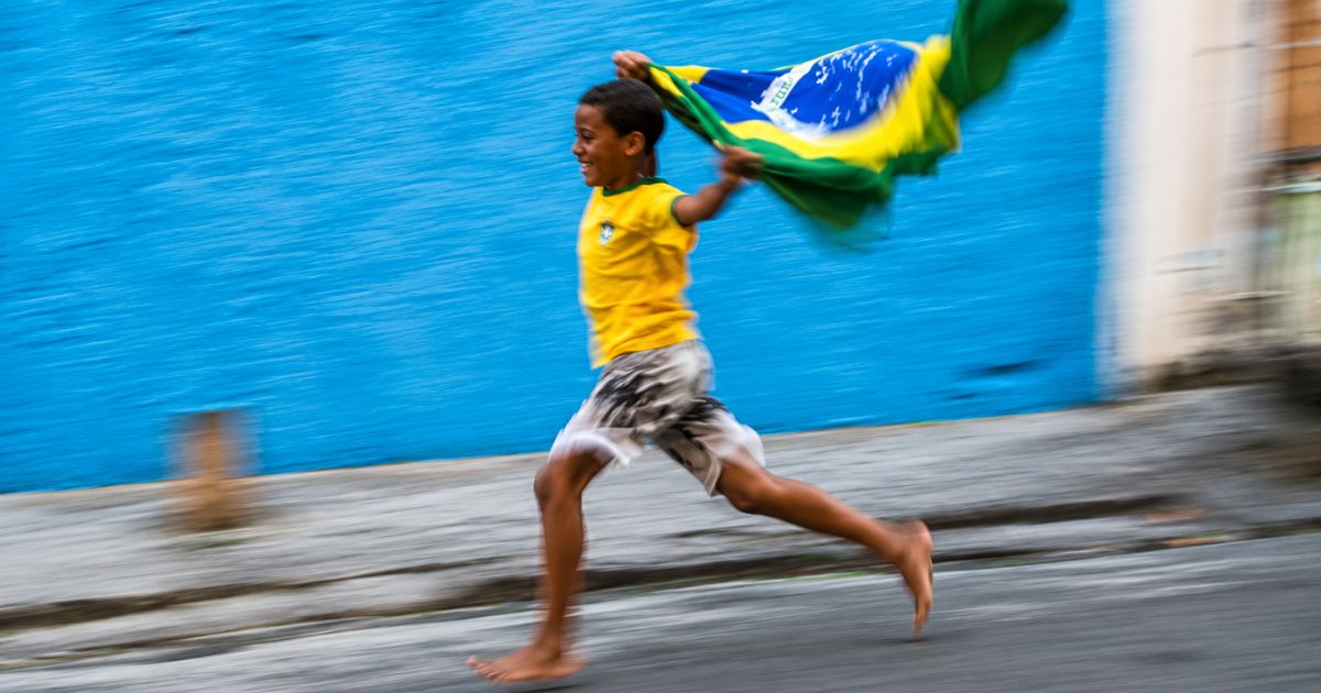 Menino sorridente, de pele morena e cabelo escuro, correndo descal&ccedil;o em rua de paralelep&iacute;pedos, vestindo camisa amarela e shorts estampados, agitando uma bandeira do Brasil. O fundo &eacute; uma parede azul vibrante, com efeito de movimento