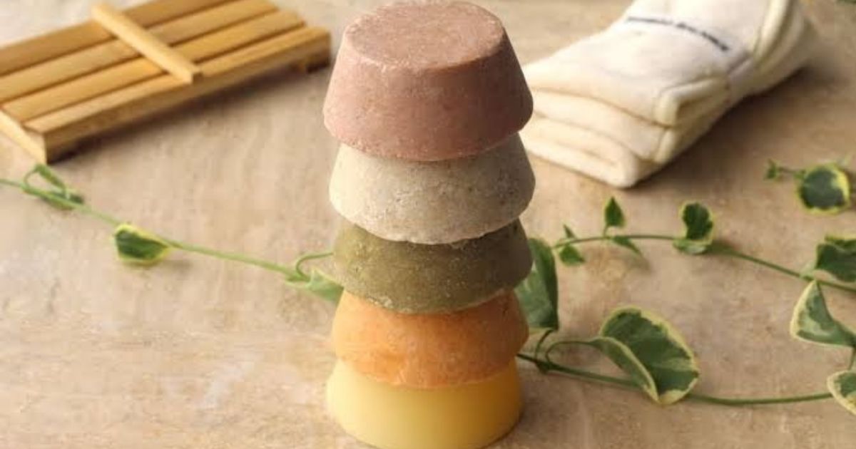 Pilha de cinco sabonetes s&oacute;lidos em formato de cone invertido, com cores que variam do rosa ao amarelo, sobre uma superf&iacute;cie clara. Ao fundo, um porta-sabonetes de madeira e uma toalha branca dobrada. Um ramo verde com folhas pequenas decora a cena