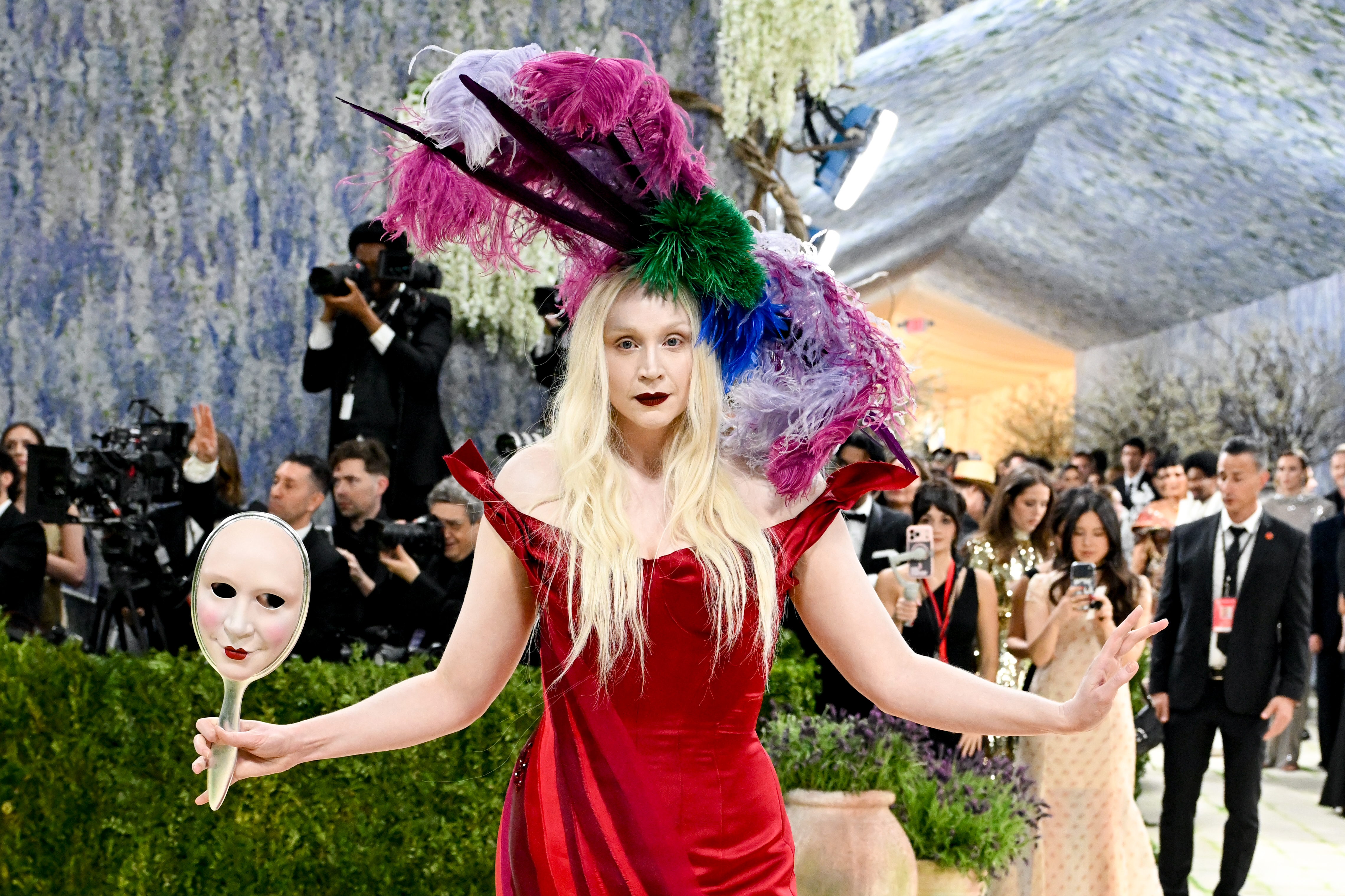 Gwendoline Christie usa vestido vermelho com headpiece exuberante e segura uma máscara no tapete vermelho do Met Gala 2026