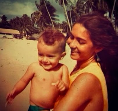 Uma mulher jovem est&aacute; segurando um beb&ecirc; no colo em uma praia ensolarada. Ela tem cabelos escuros, presos para tr&aacute;s, e est&aacute; sorrindo enquanto olha para a crian&ccedil;a. Veste uma blusa clara sem mangas. O beb&ecirc;, que parece ter cerca de um ano, est&aacute; sem camisa, usando apenas um short, e tamb&eacute;m sorri, olhando levemente para frente.