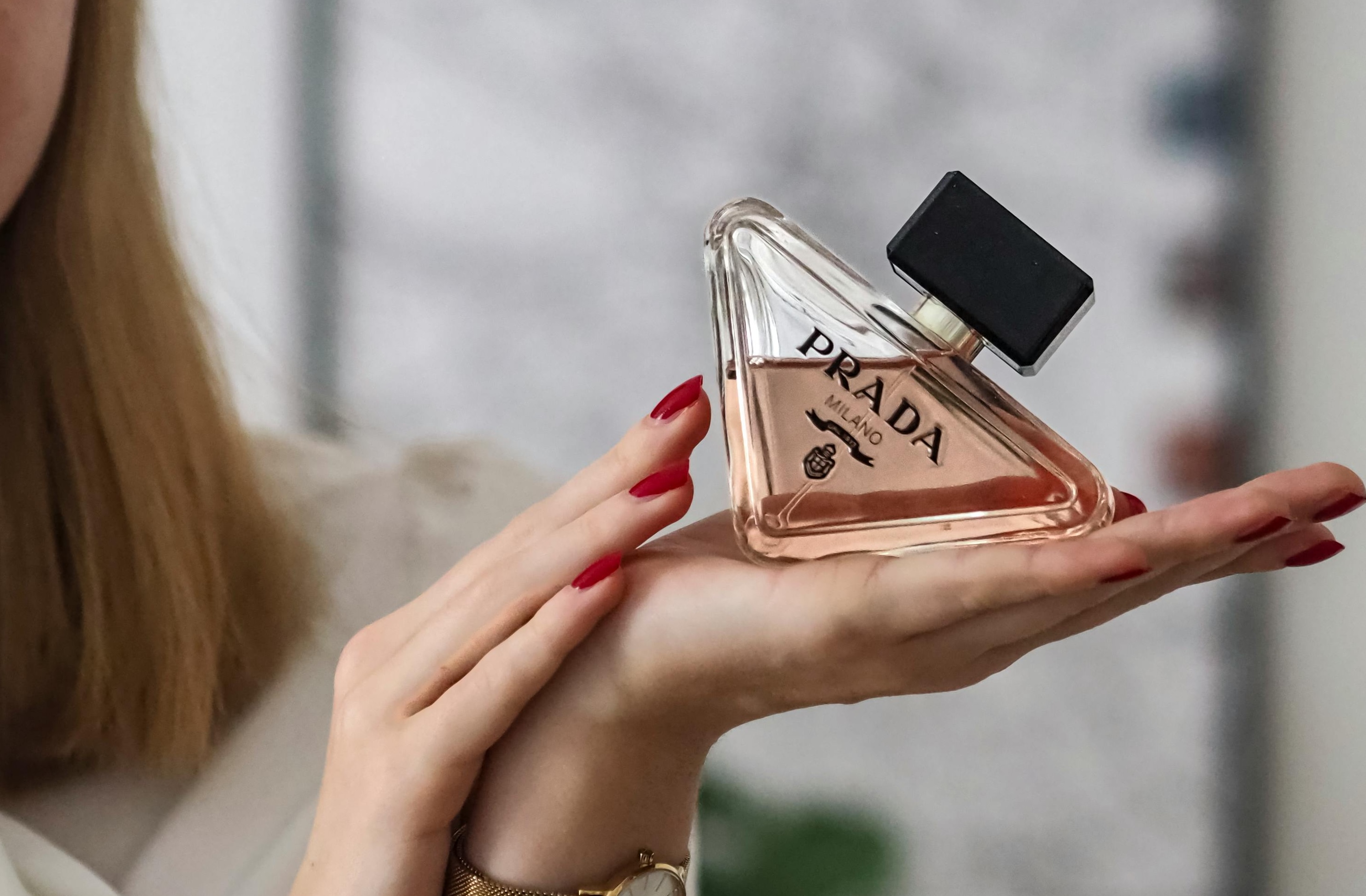 Mão feminina com unhas vermelhas segurando um frasco de perfume triangular da marca Prada Milano, com líquido rosado e tampa preta. Um relógio dourado é visível no pulso.