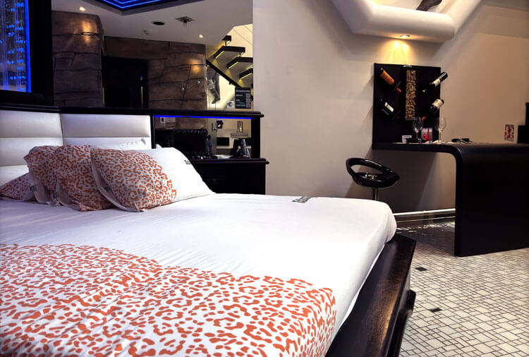 Quarto de motel com cama de casal, len&ccedil;&oacute;is brancos e almofadas com estampa de on&ccedil;a laranja. Ao fundo, balc&atilde;o preto com garrafas de vinho, ta&ccedil;as e um banco alto.