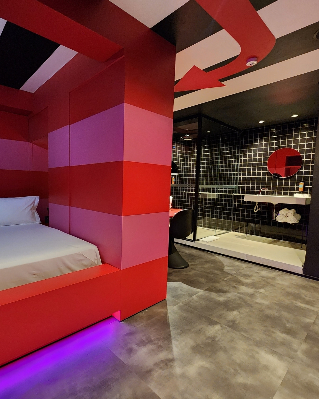 Quarto moderno com cama vermelha e rosa, ilumina&ccedil;&atilde;o roxa no rodap&eacute;, e banheiro integrado com paredes de azulejos pretos e brancos, pia branca e espelho redondo vermelho. O teto tem listras pretas e brancas com uma seta vermelha