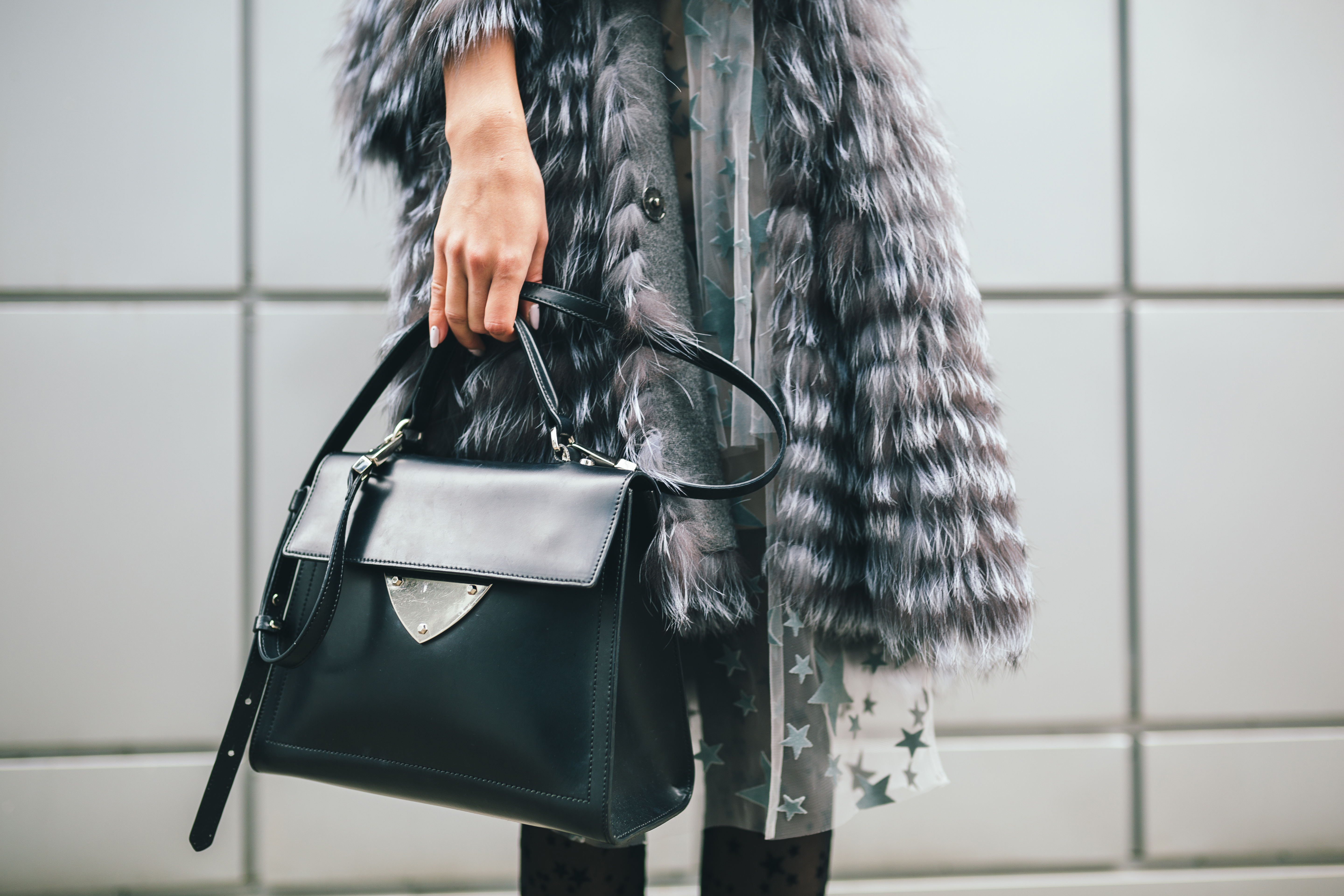 Close de uma bolsa preta de luxo sendo segurada por uma mulher usando casaco de pele sint&eacute;tica cinza e vestido transparente com estampas de estrelas.
