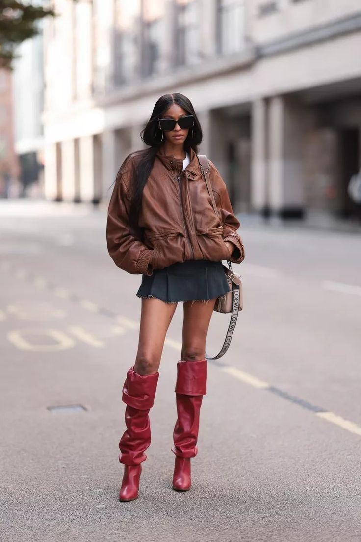 Modelo posa no meio da rua usando jaqueta bomber marrom, minissaia preta e botas slouchy vermelhas com cano franzido.