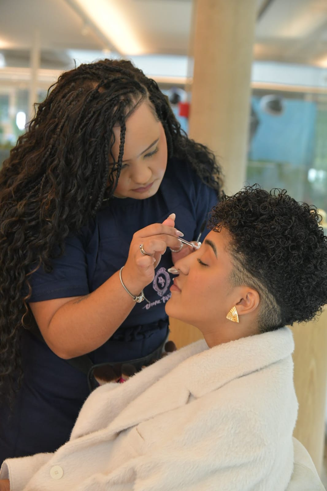 Maquiadora de pele clara e cabelos longos em tran&ccedil;as, vestindo camiseta azul, aplica maquiagem nos olhos fechados de uma mulher de pele morena, cabelo crespo curto com laterais raspadas e brinco dourado triangular, usando um roup&atilde;o branco