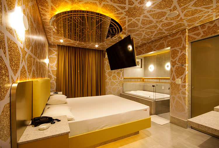 Quarto de motel luxuoso com paredes e teto dourados, cama de casal branca com cabeceira dourada, TV preta suspensa, banheira de hidromassagem e cortinas douradas