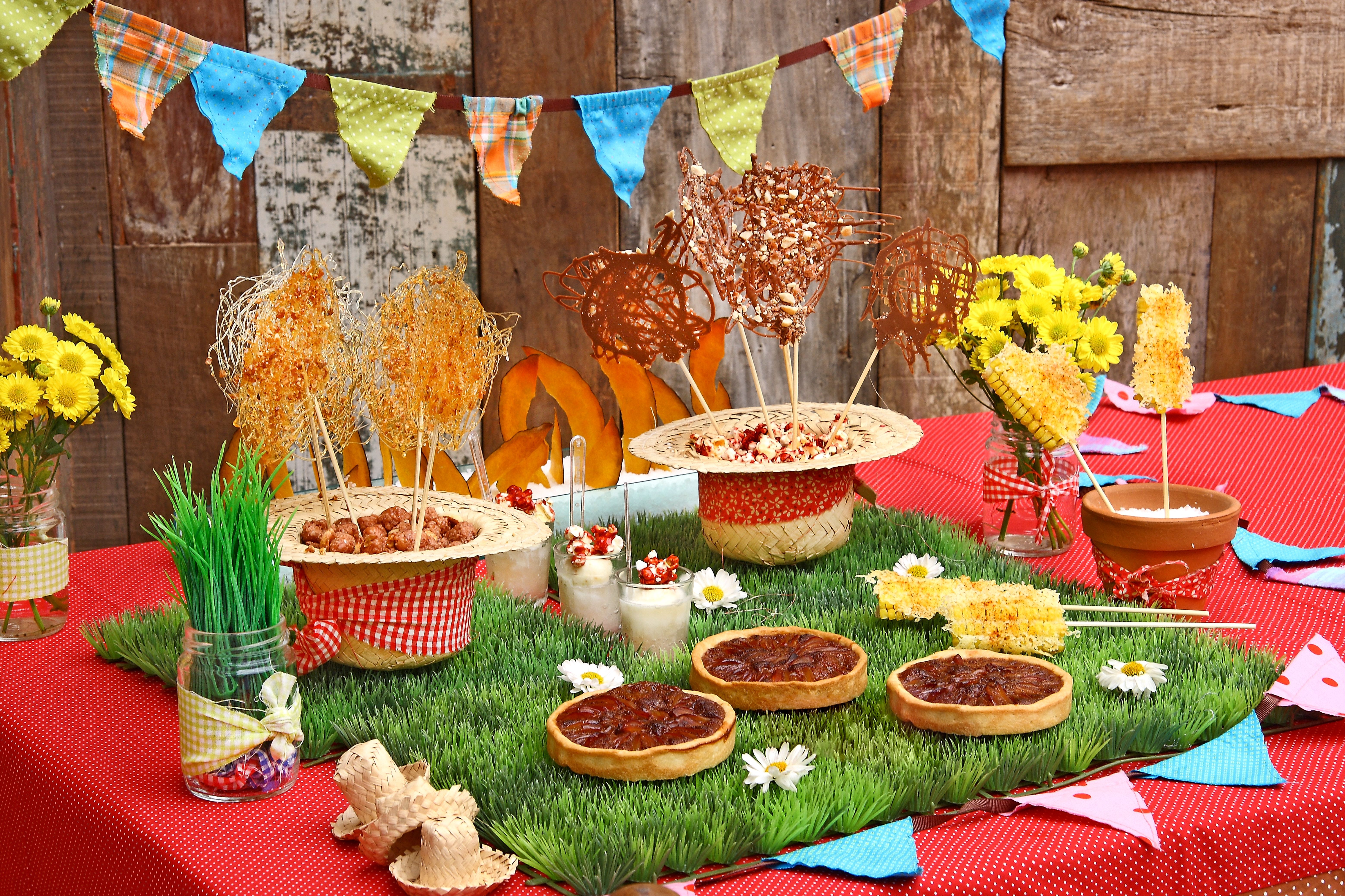 Mesa de festa junina com toalha vermelha, grama sintética, bandeirinhas coloridas, doces em espetos e tortinhas, milho, flores amarelas e chapéus de palha