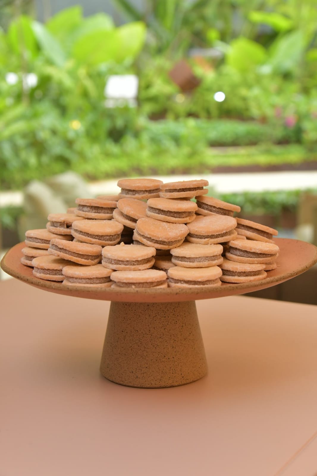 Pequenos biscoitos redondos com recheio escuro, empilhados em um prato de cer&acirc;mica marrom sobre uma mesa rosa, com folhagem verde desfocada ao fundo